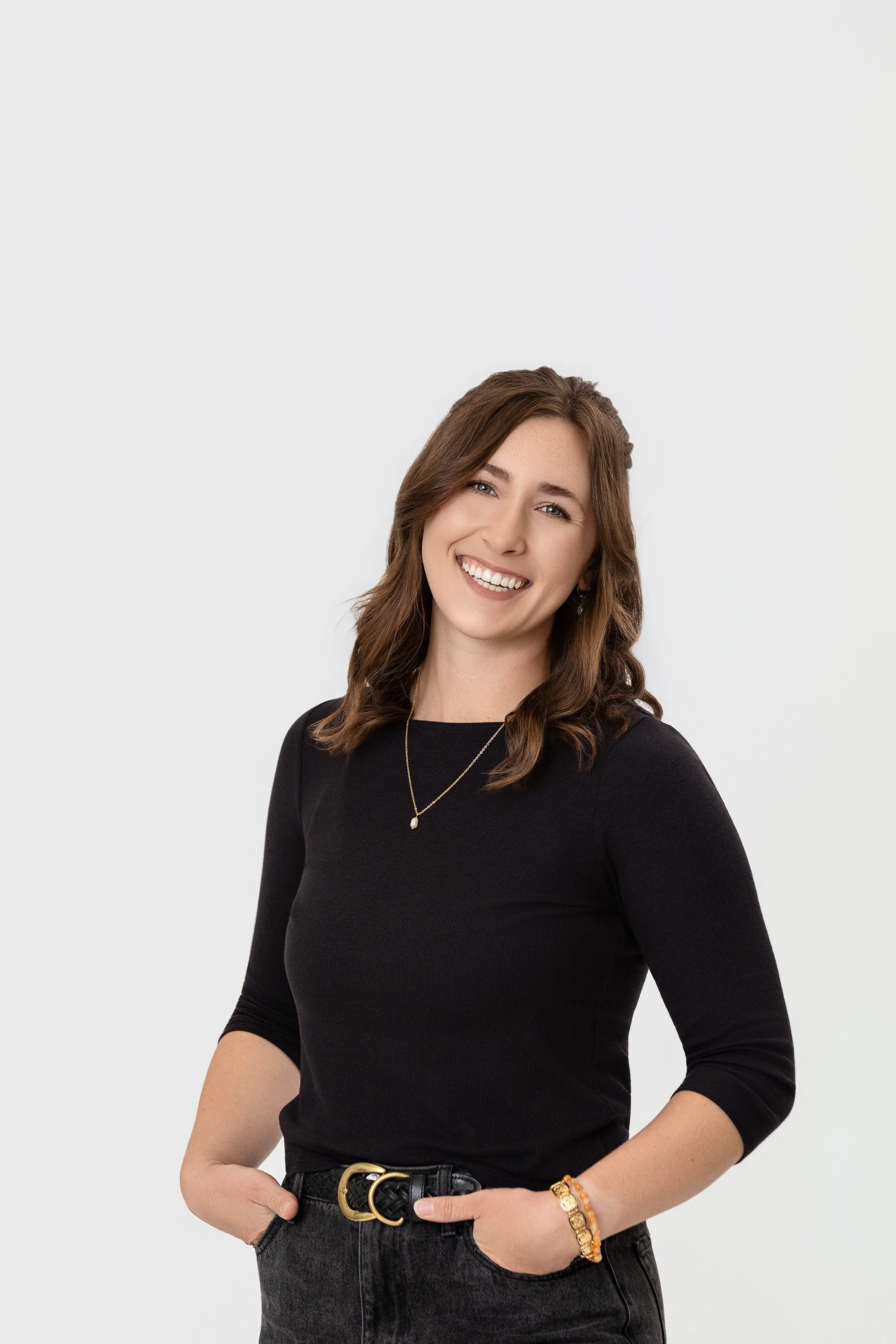 Color portrait of a smiling woman with long wavy brown hair, standing and smiling with hands in pockets