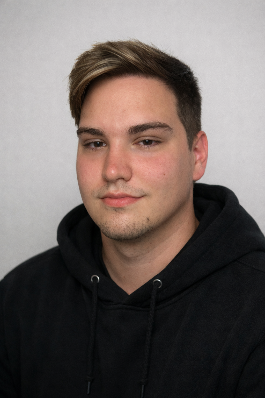A young man with brown hair and light skin, wearing a black hoodie, looking at the camera with a slight smile, against a plain light gray background.