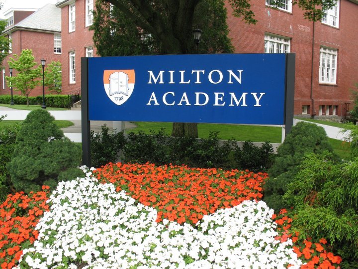 Milton Academy sign with shield logo, surrounded by orange, white, and green flowers and greenery, in front of a brick building and trees.