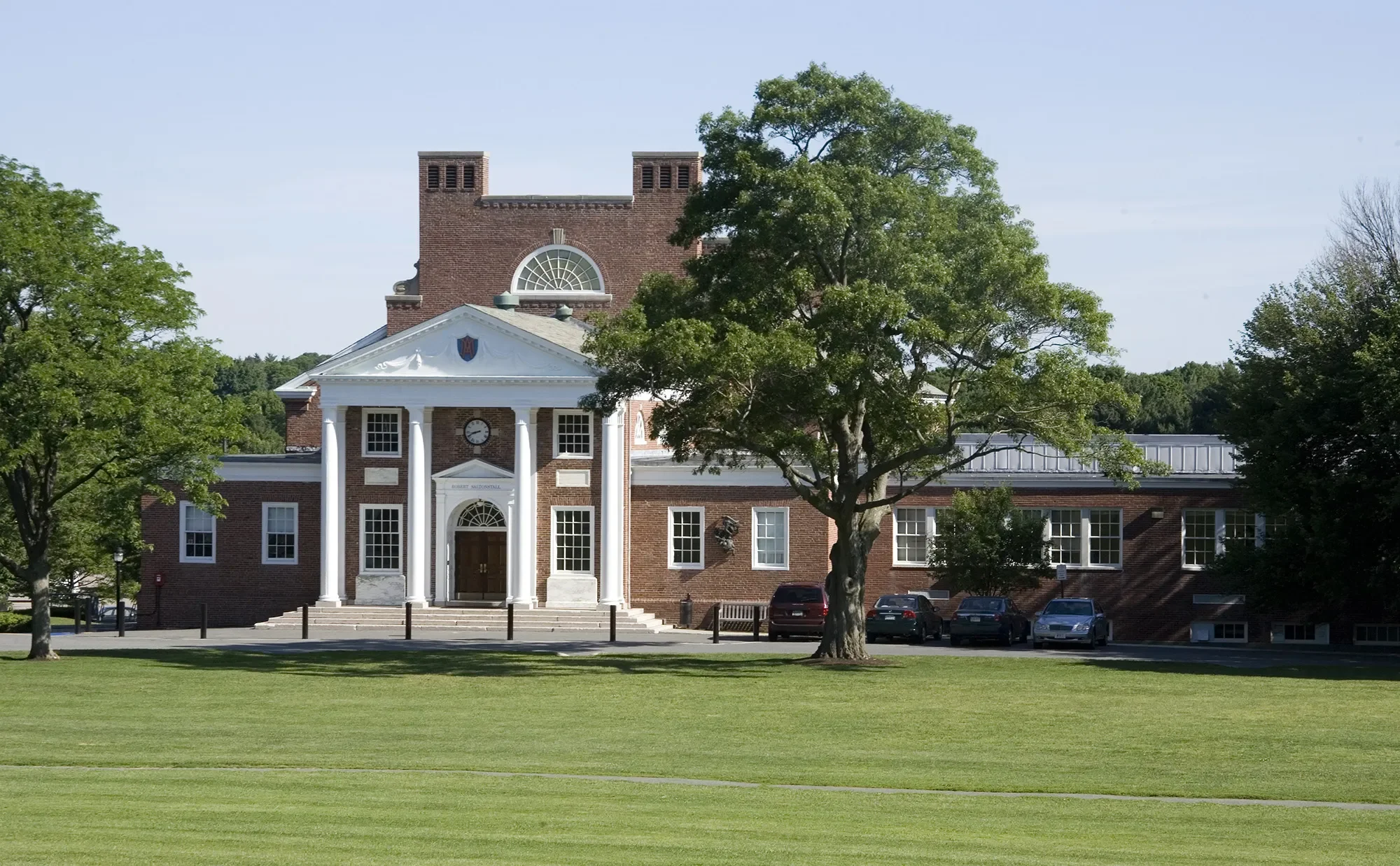 A university building with a central entrance featuring four white columns, a brick facade, a clock above the entrance, and surrounding trees on a well-maintained grassy lawn.