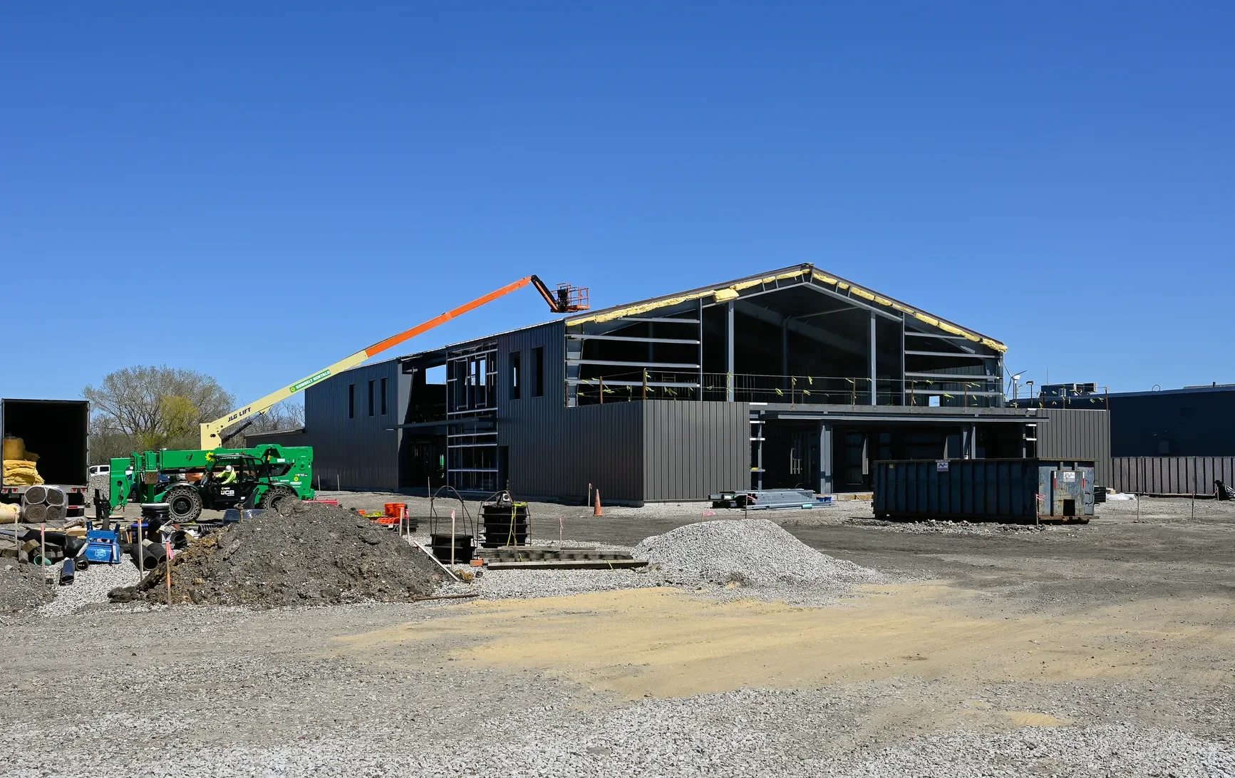 Construction site with a partly built black metal building, construction equipment, piles of dirt and gravel, and a clear blue sky.