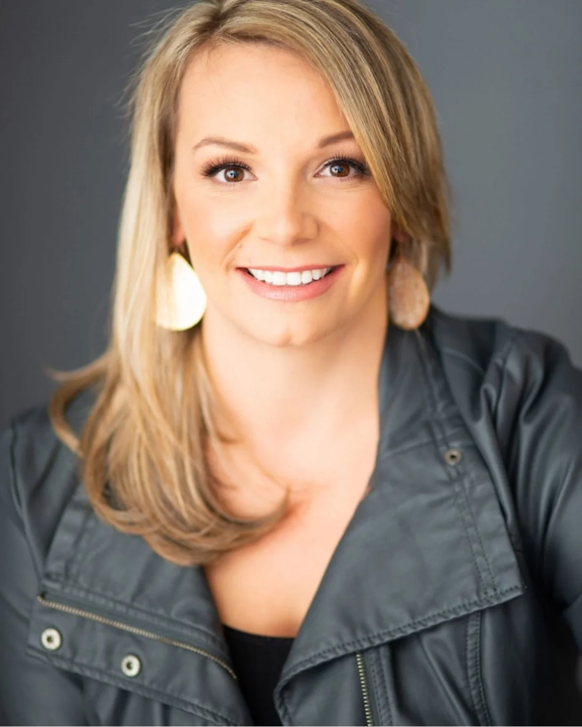 Portrait of a smiling woman with blond hair wearing a black leather jacket and beige earrings against a gray background.