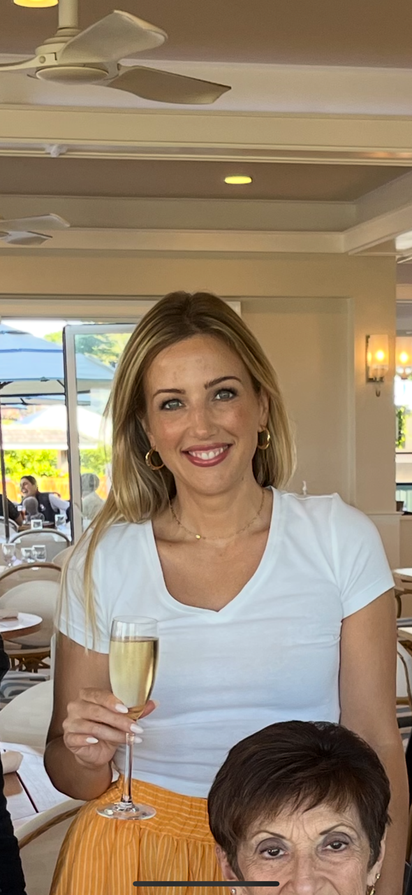 A woman with blonde hair holding a glass of champagne, smiling in a bright restaurant or cafe setting with people in the background.