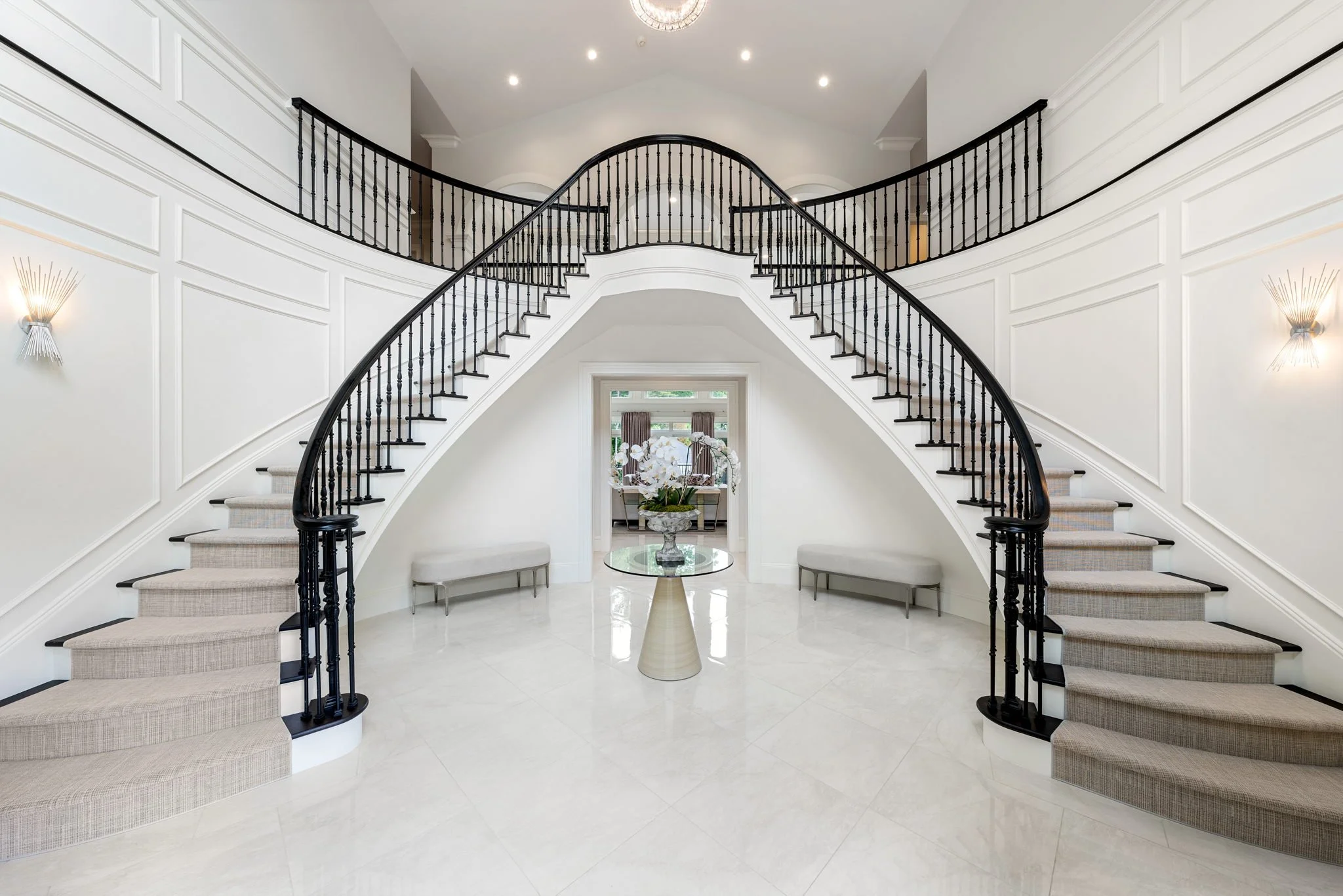 Elegant foyer with a grand curved staircase with black railings, white walls with decorative paneling, wall sconces, and a large glass-top table with a floral arrangement in the center.