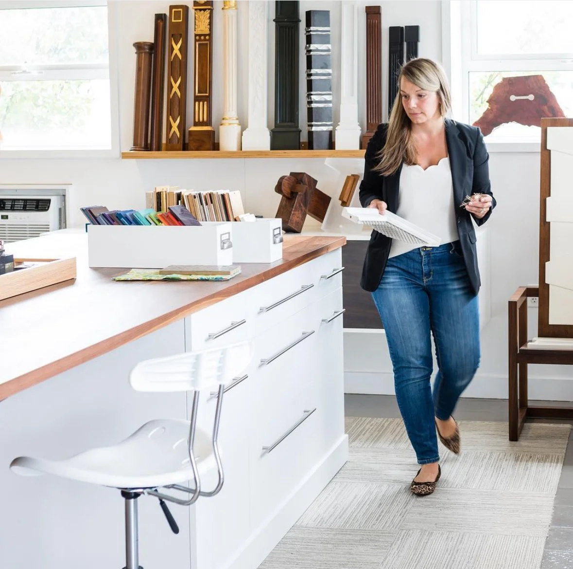 Woman in a blazer and jeans walking in a modern office or studio space with wooden and white furniture, books, and decorative wood pieces on the shelves.