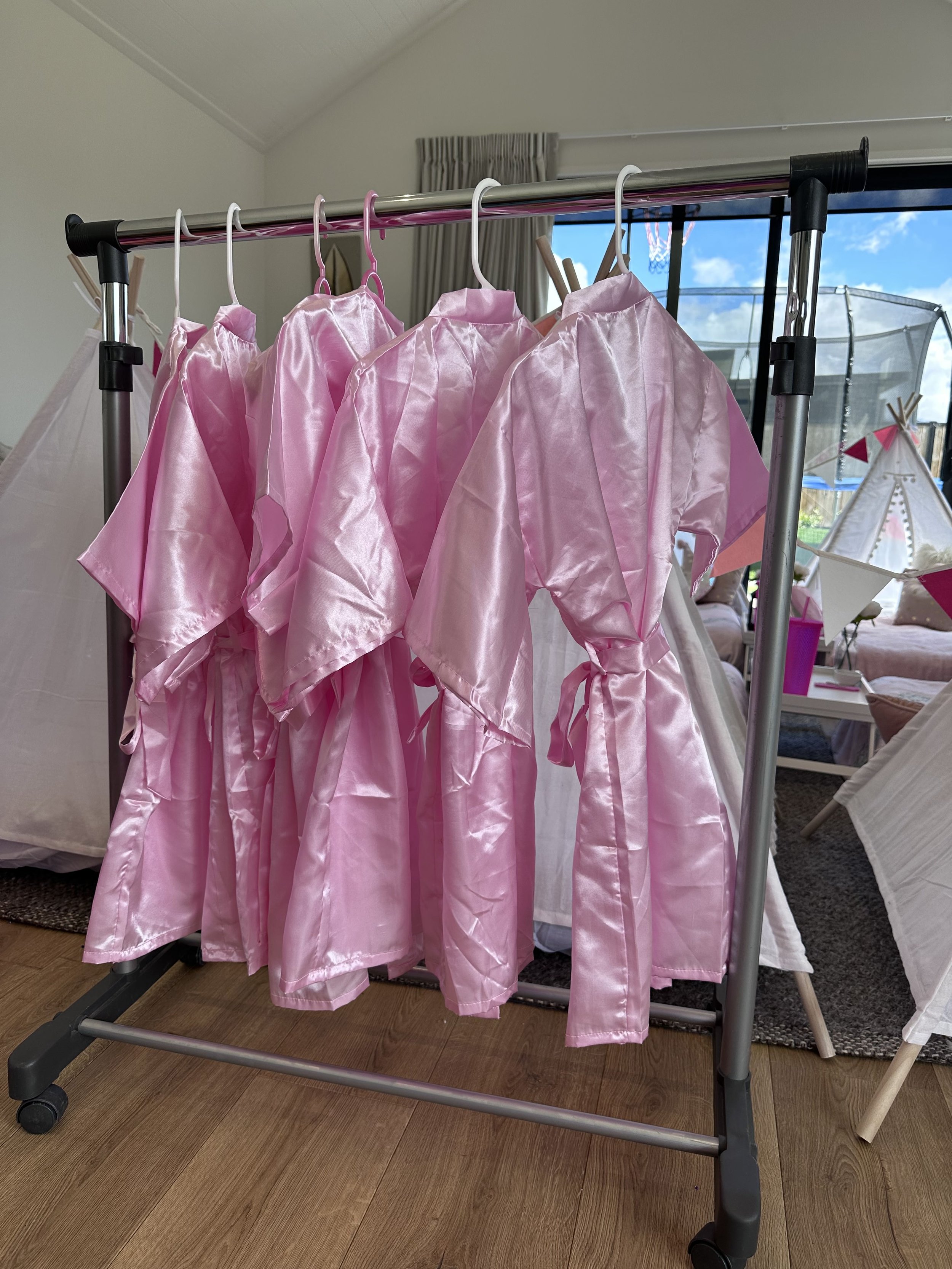 A clothing rack with several pink satin robes hanging on hangers, ready for SPA Girls Night slumber party.
