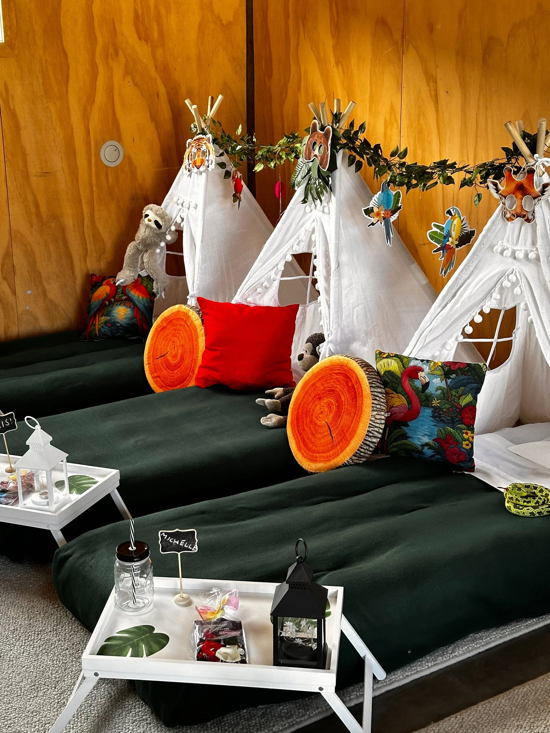 Plush animals and animal-themed pillowcases decorate the beds, a garland with animal cutouts hangs, and a small table with treats and a lantern is placed in front of the beds. Rainforest ready setup for slumber party.
