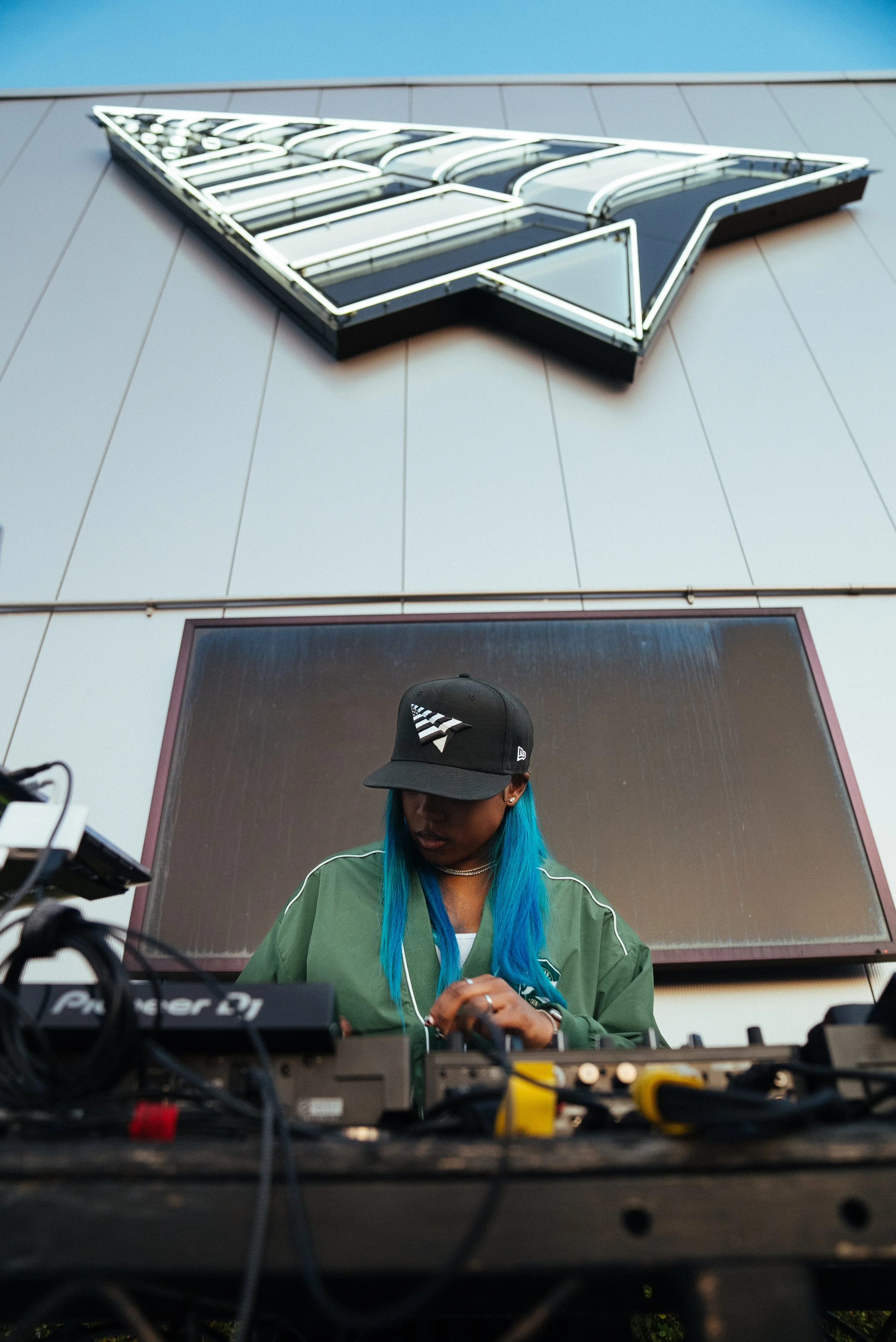 Sounds Of Reality At Planes - A woman with blue hair and a black cap DJing at an outdoor event with a large building and a neon sign in the background.