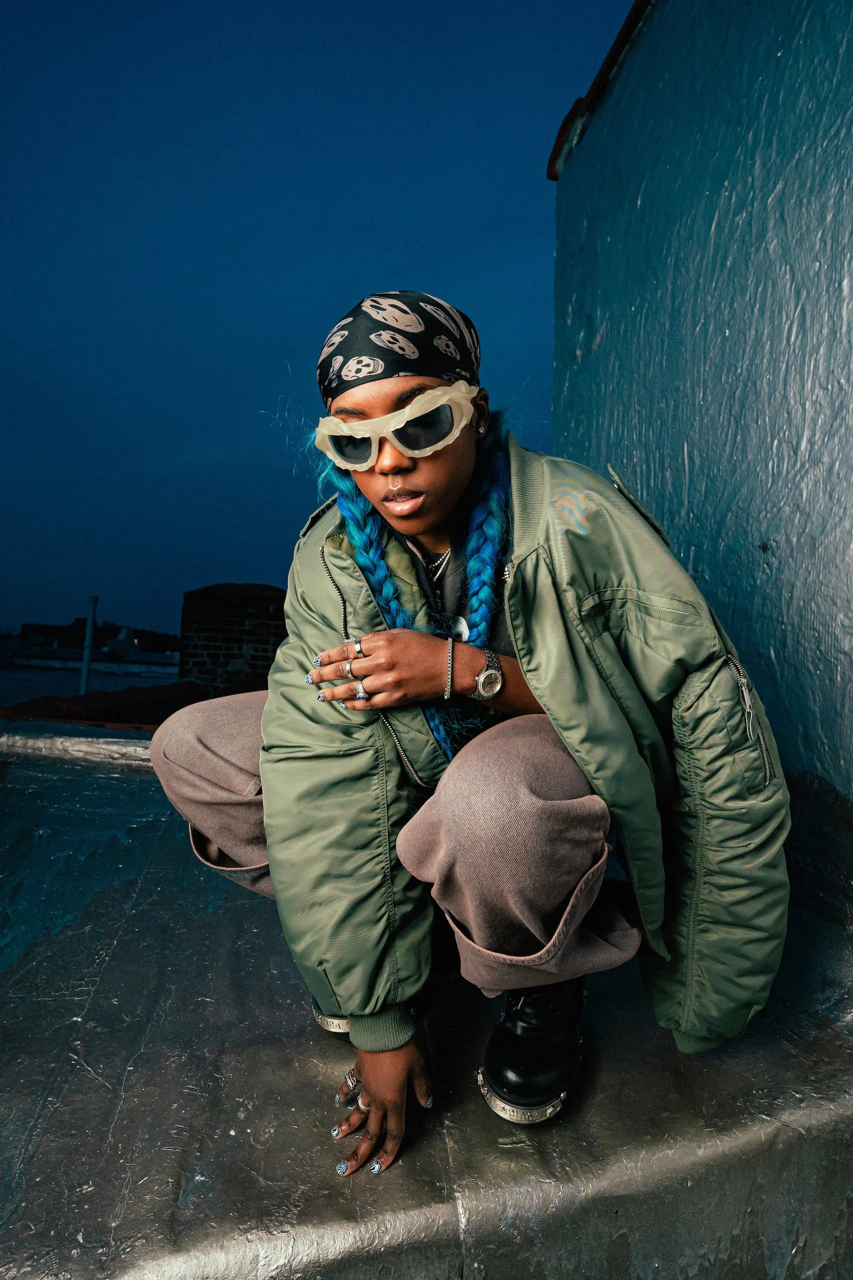 Person squatting on a rooftop at dusk wearing sunglasses, a bandana, green jacket, and cargo pants with bold nails, multiple rings, and a watch.