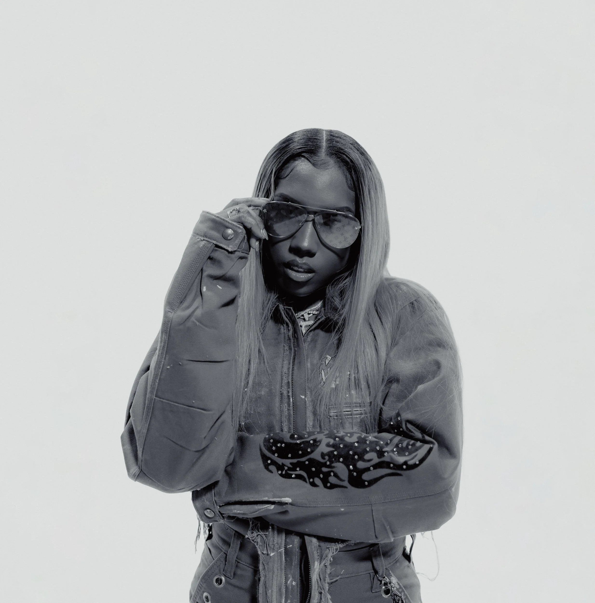 Black and white photo of a woman wearing sunglasses and a jacket, looking at the camera with one hand adjusting her sunglasses, against a plain light background.