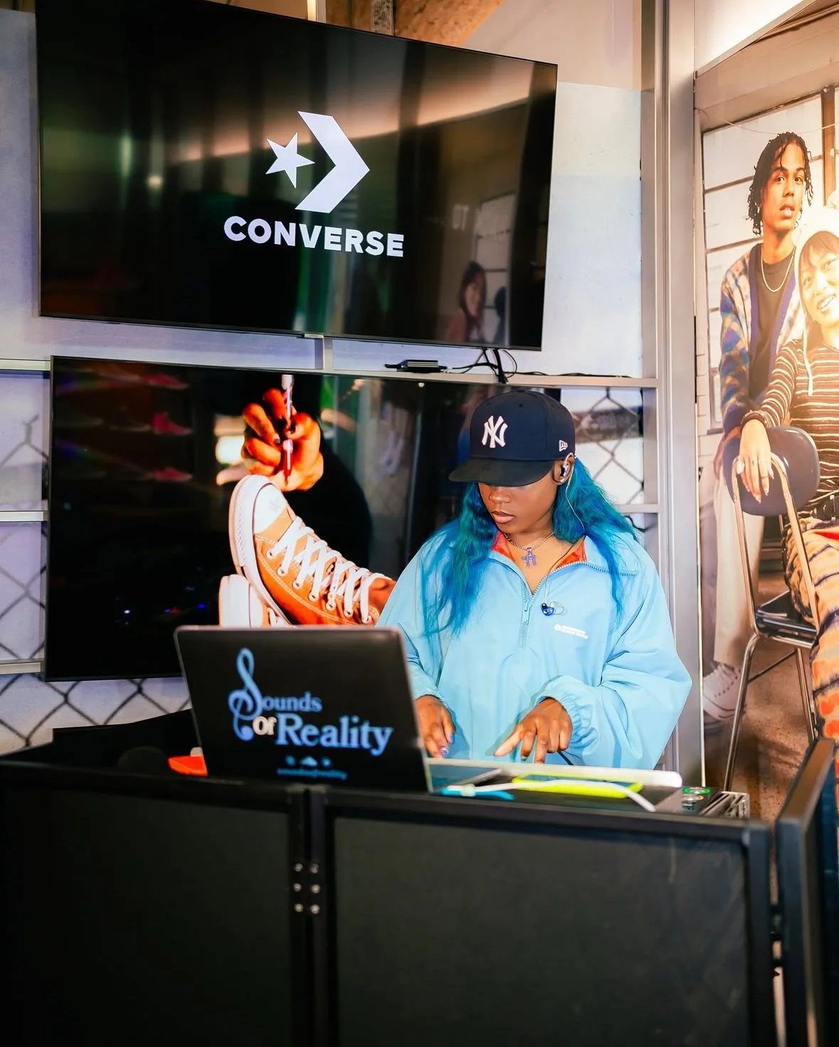 A woman with blue hair wearing a New York Yankees cap and light blue jacket is DJ-ing at a booth, with a laptop that reads 'Sounds of Reality', in front of a large television screen displaying the Converse logo. The background features a poster of tw