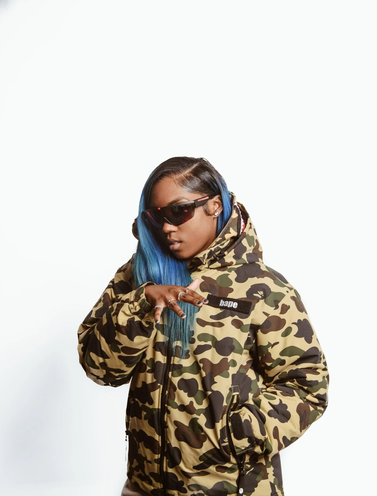 A woman with blue hair wearing a camouflage jacket and sunglasses, posing with her hand extended in a peace sign.