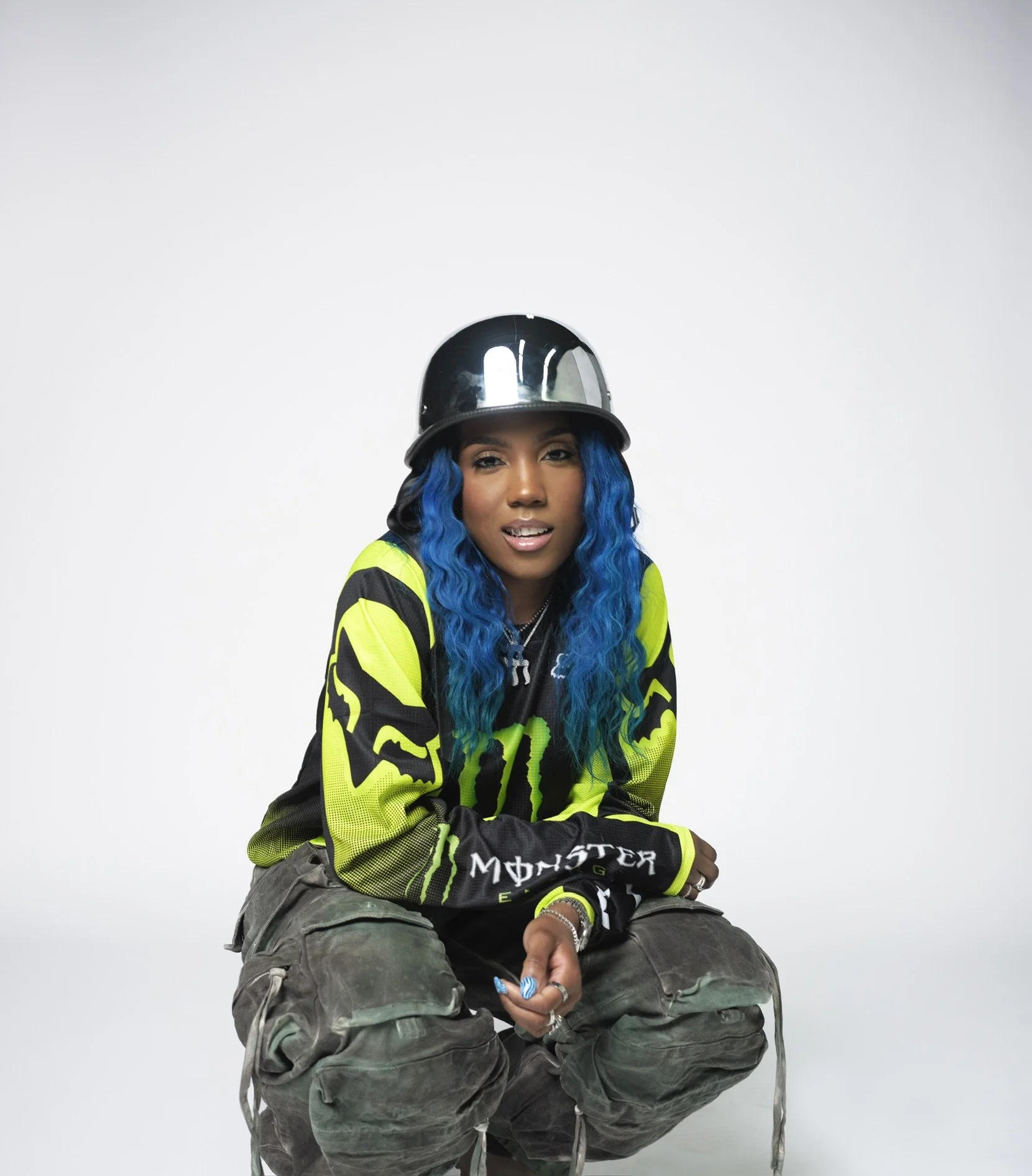 A woman with long, wavy blue hair wearing a black helmet, a black and neon yellow Monster Energy racing jacket, and camouflage pants, sitting against a white background.