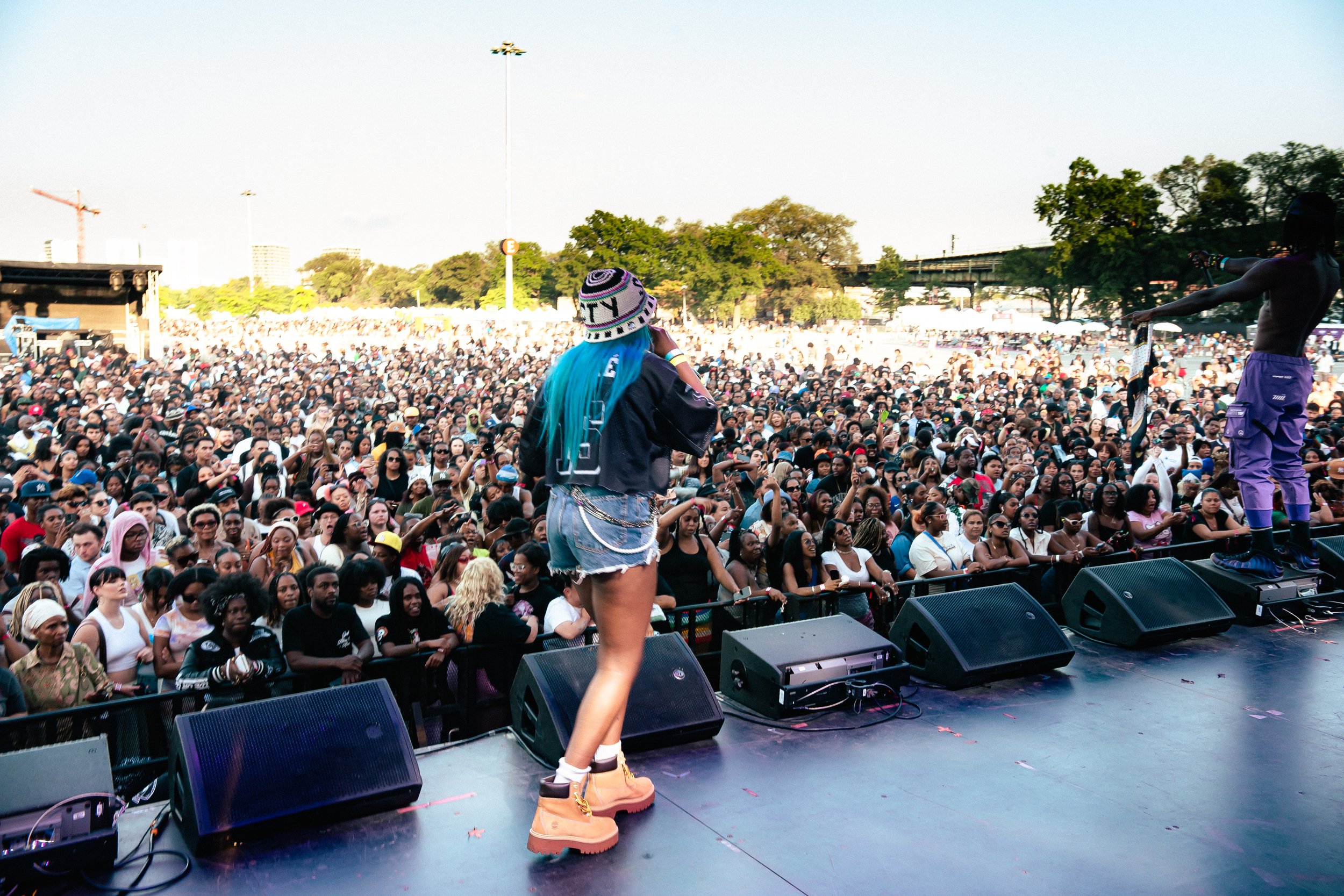 Sounds Of Reality Performer with bright blue hair and tan boots on stage at outdoor music concert, facing large crowd of people.