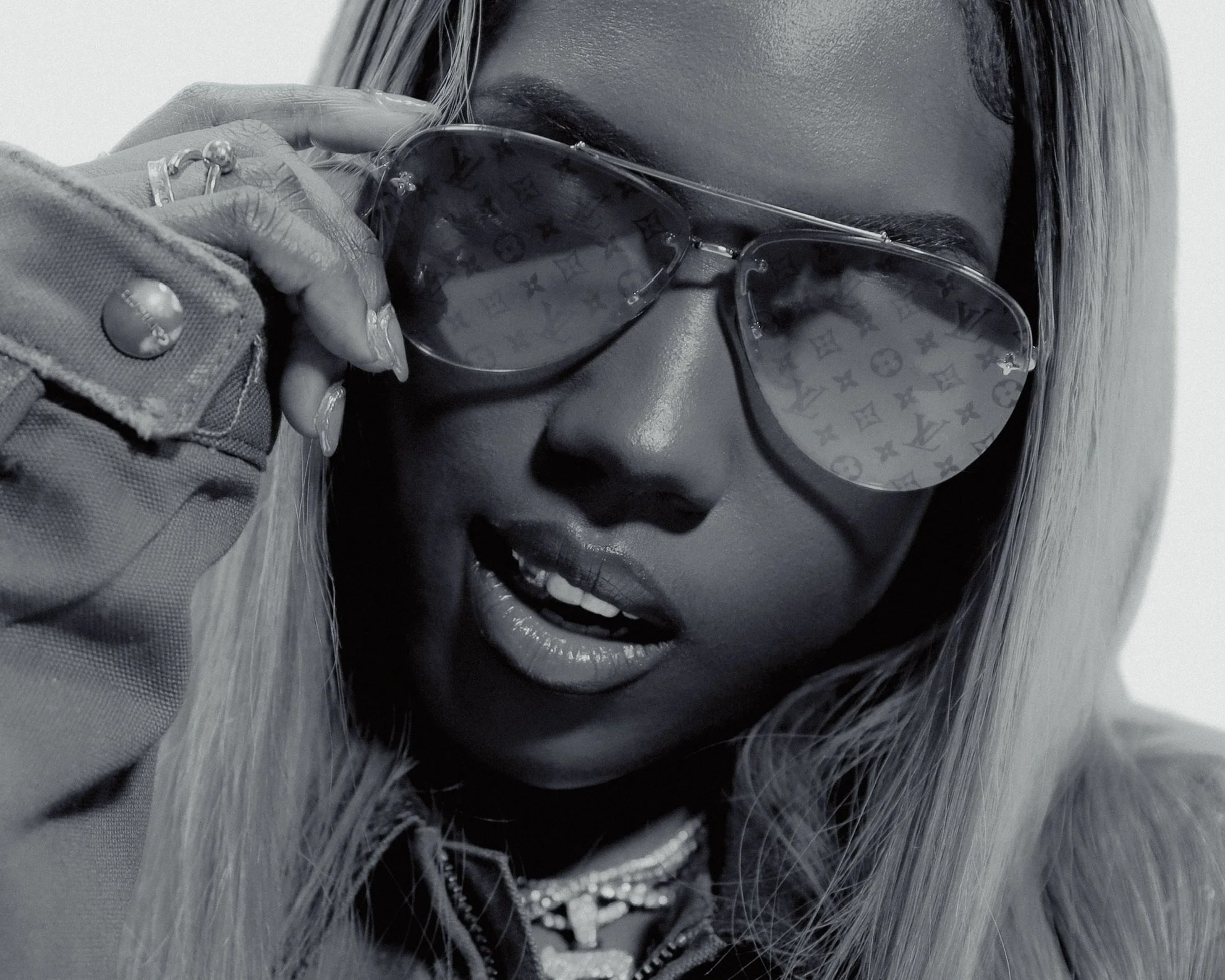 A close-up black and white photo of a woman wearing sunglasses with Louis Vuitton pattern, holding the frame with her hand, and slightly opening her mouth to reveal her teeth.