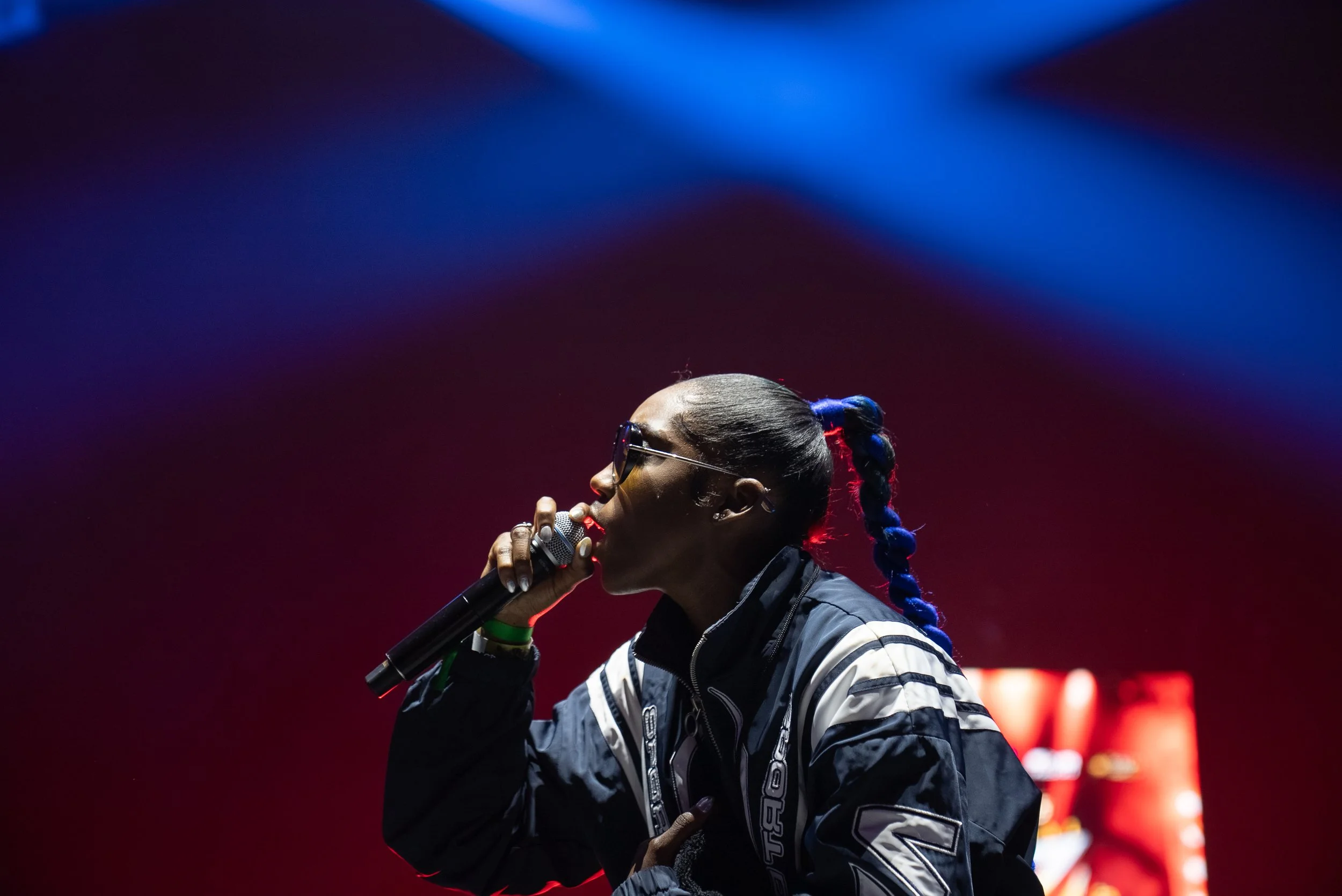 A woman with braided hair and sunglasses singing into a microphone on a stage with colorful stage lights.