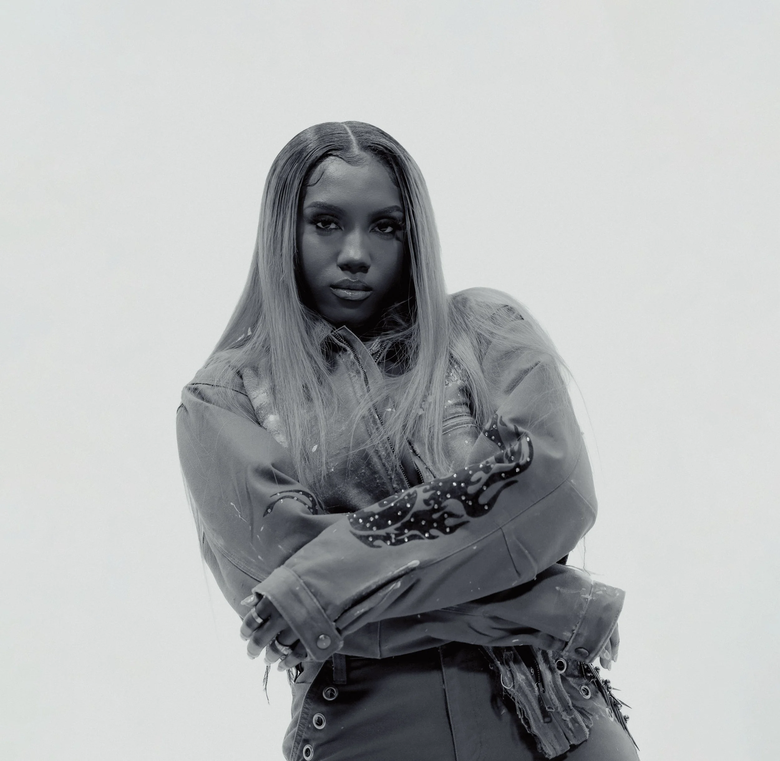 Black woman with long hair wearing a leather jacket with a flaming design on the sleeve, crossing her arms, against a plain light background.