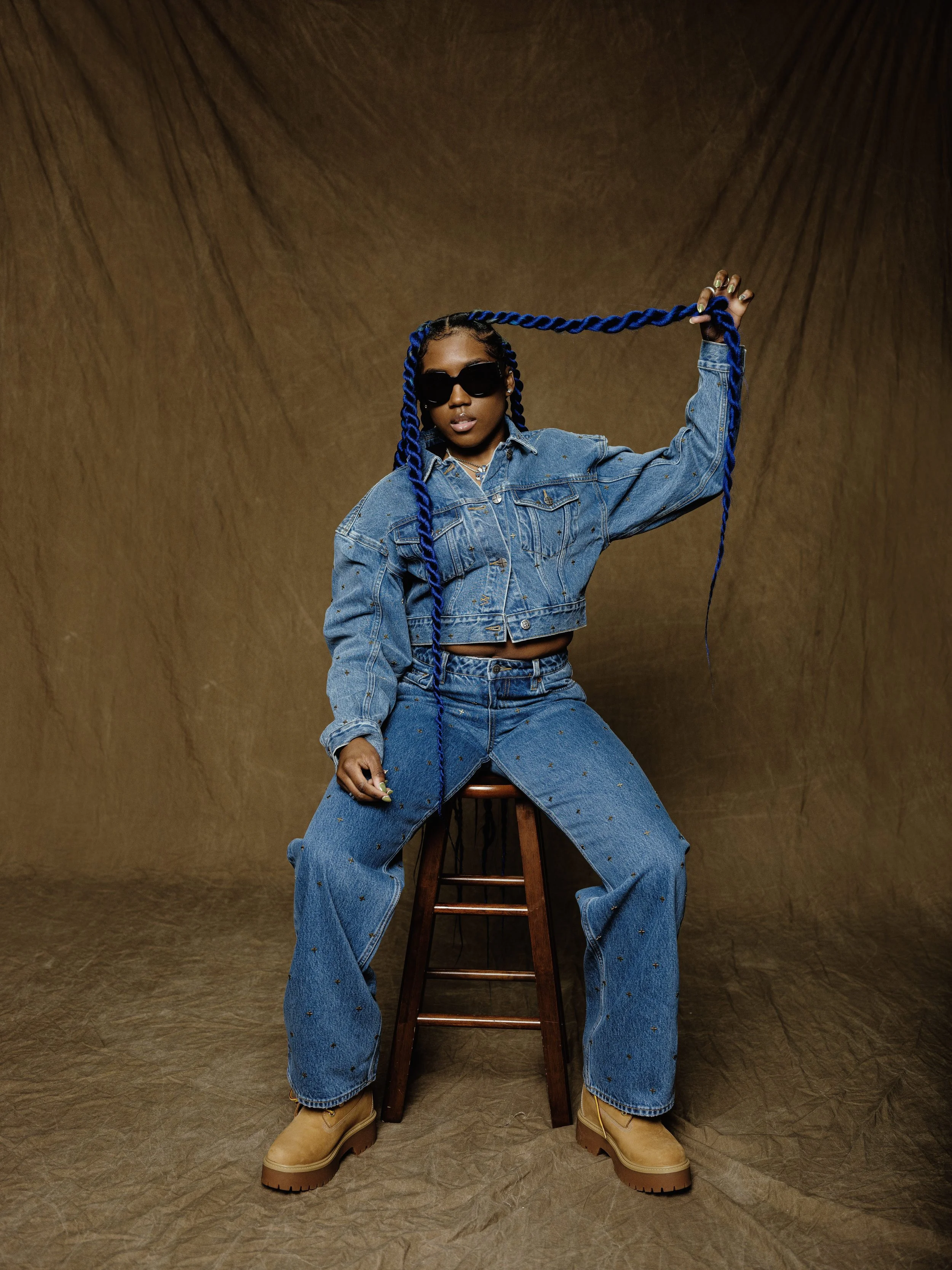 Sounds Of Reality with long braided blue hair, wearing sunglasses, a denim jacket, and matching jeans, sitting on a wooden stool against a brown backdrop, holding her braided hair with one hand.