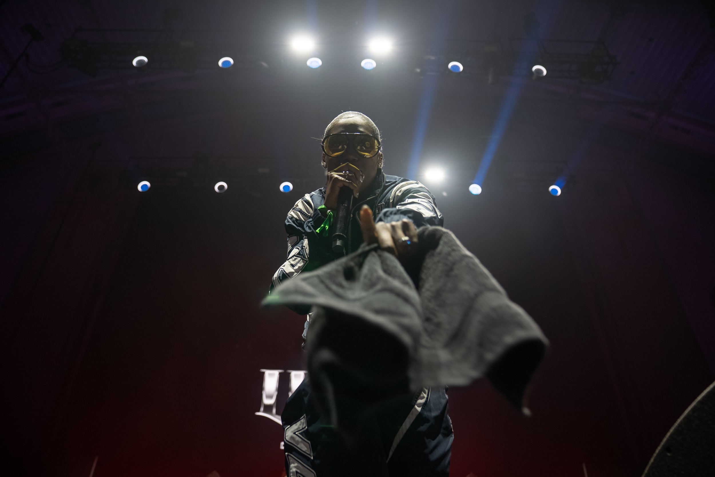 Performer on stage during concert, wearing sunglasses, holding microphone, reaching towards camera, backlit with stage lights.