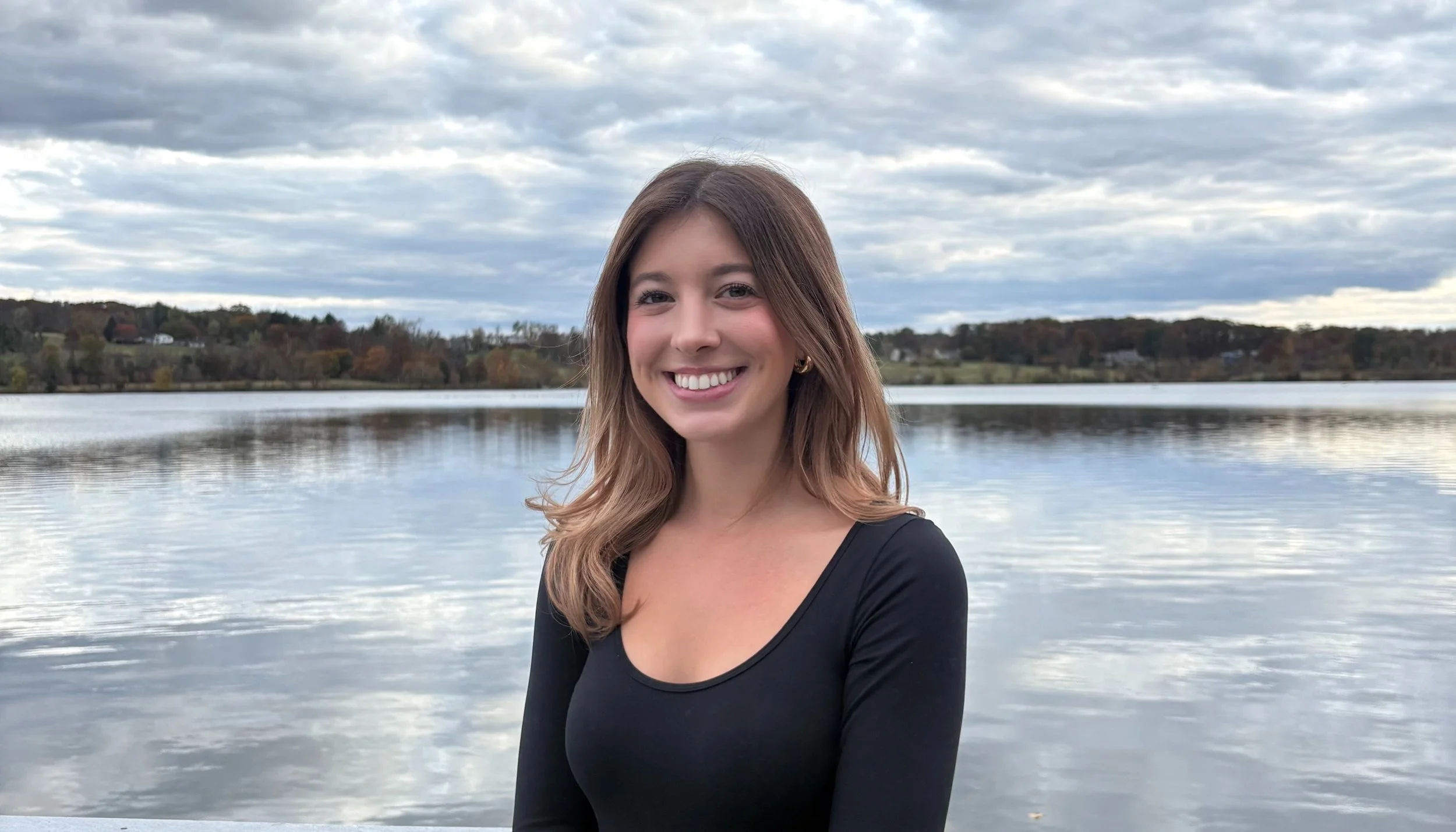A young woman with shoulder-length brown hair smiling at the camera, wearing a black top, standing by a calm lake with hills and houses in the background under cloudy sky.