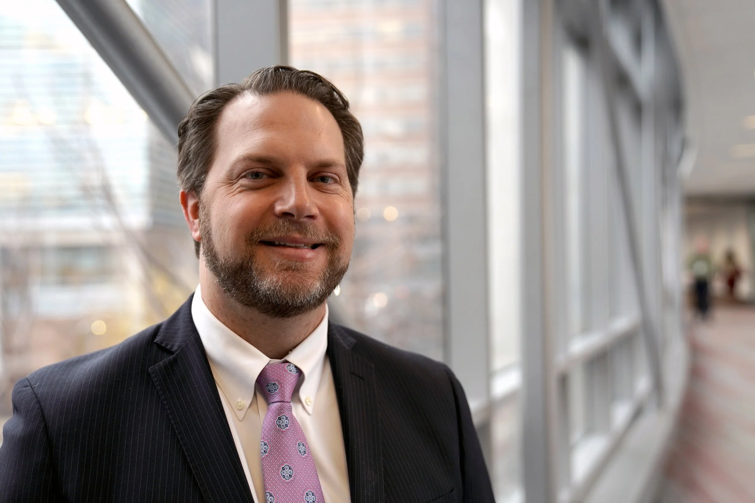 A man with brown hair and a beard, wearing a dark suit, white shirt, and light purple tie with small patterns, smiling while standing indoors near large windows with a cityscape in the background.