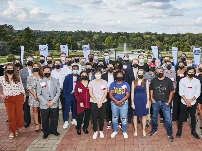 Group of people wearing masks standing outdoors on a brick pathway, with banners and trees in the background.