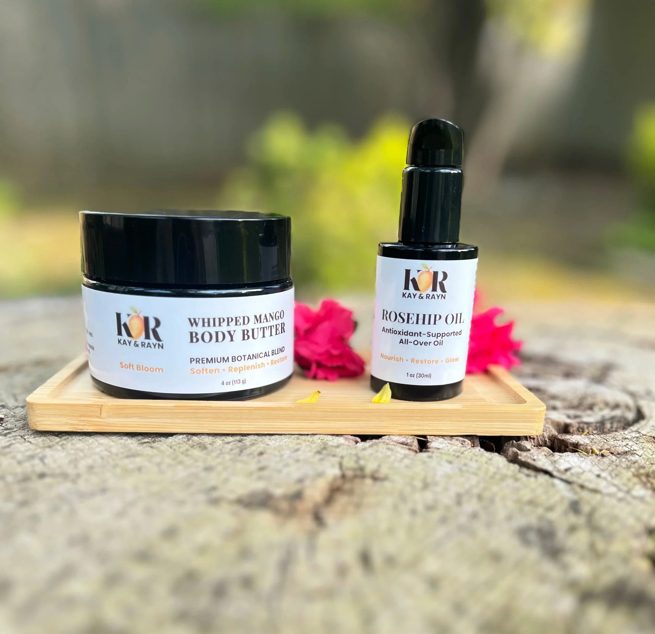 Body care duo - body butter and rosehip oil, two black containers of Kay & Rayn skincare products placed on a wooden tray with pink flower petals in the background, outdoors.