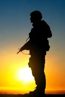 Silhouette of a person with a backpack holding a rifle, standing outdoors during sunset or sunrise.