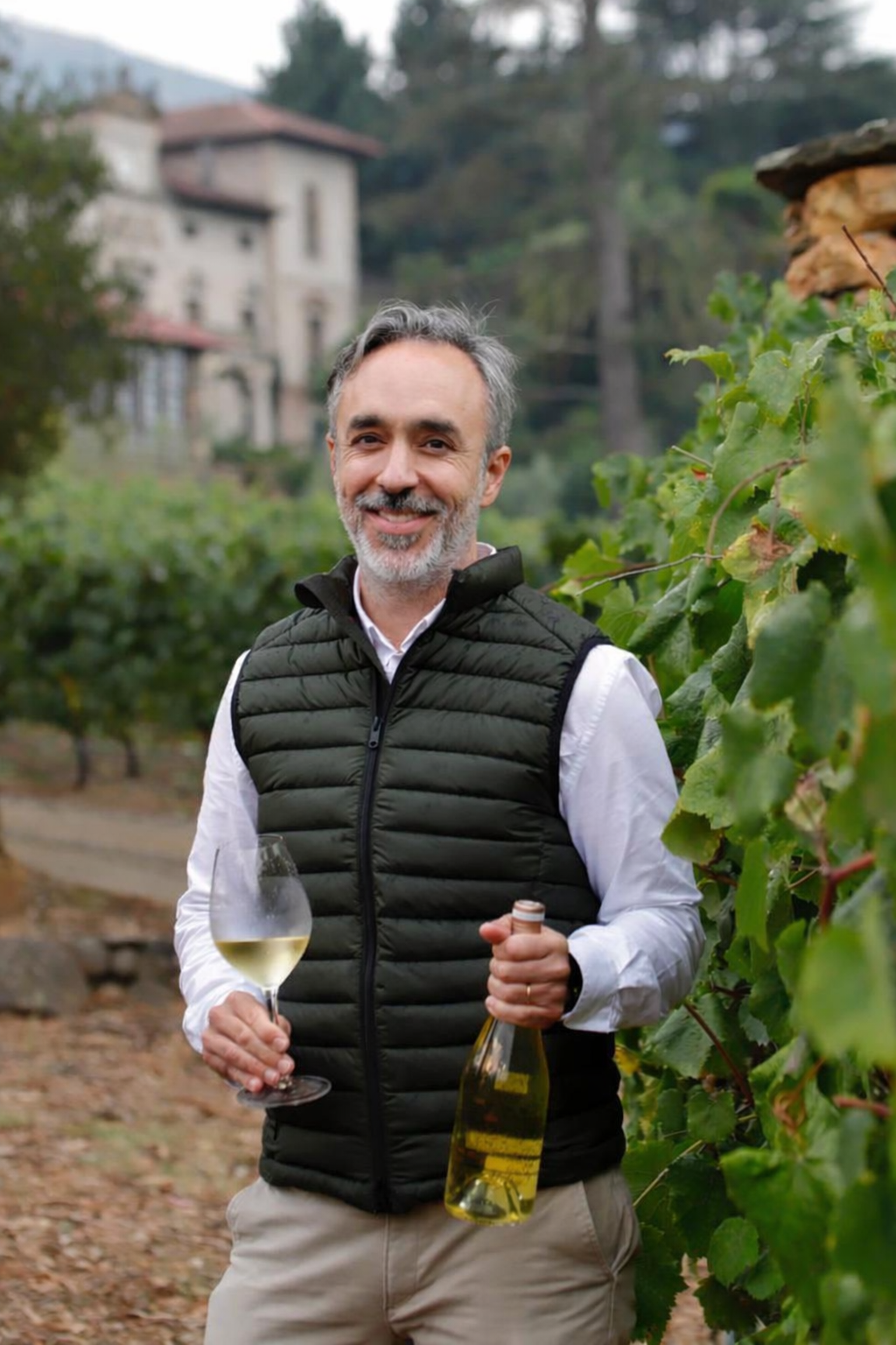 Hombre sonriendo en un viñedo, sosteniendo una copa de vino blanco y una botella de vino.
