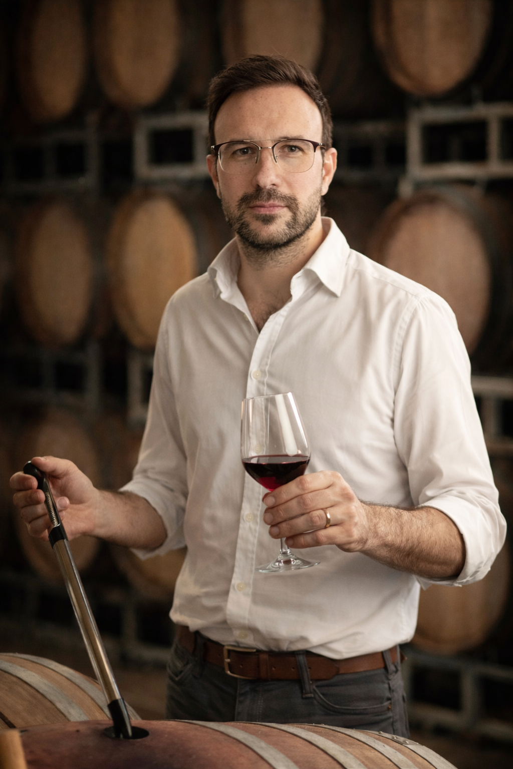 Hombre con barba y gafas, vestido con camisa blanca, sosteniendo una copa de vino tinto en un viñedo con barriles en el fondo.