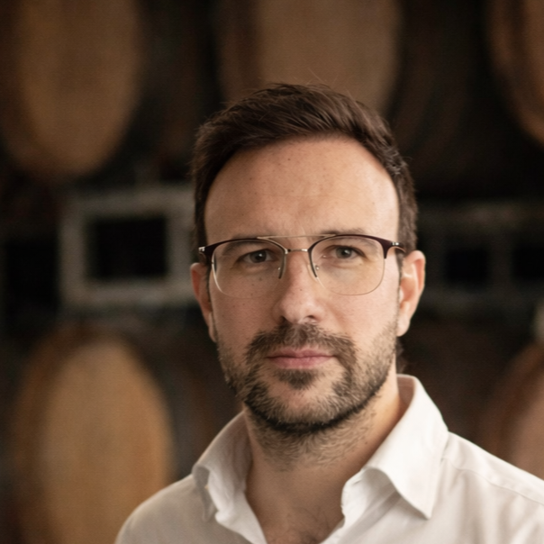Retrato de un hombre con gafas, barba y cabello castaño oscuro, con fondo de barriles de madera.
