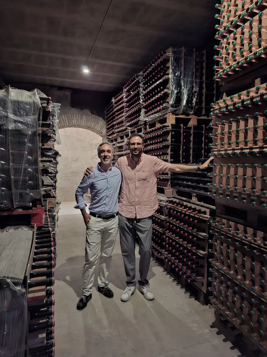Dos hombres sonrientes en una bodega con estantes llenos de botellas de vino almacenadas en palets de madera.