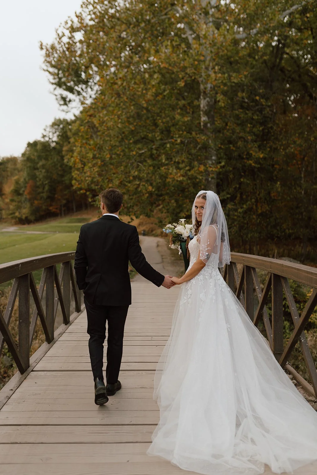 A bride and groom holding hands on a wooden bridge in a park, with trees and autumn foliage in the background.