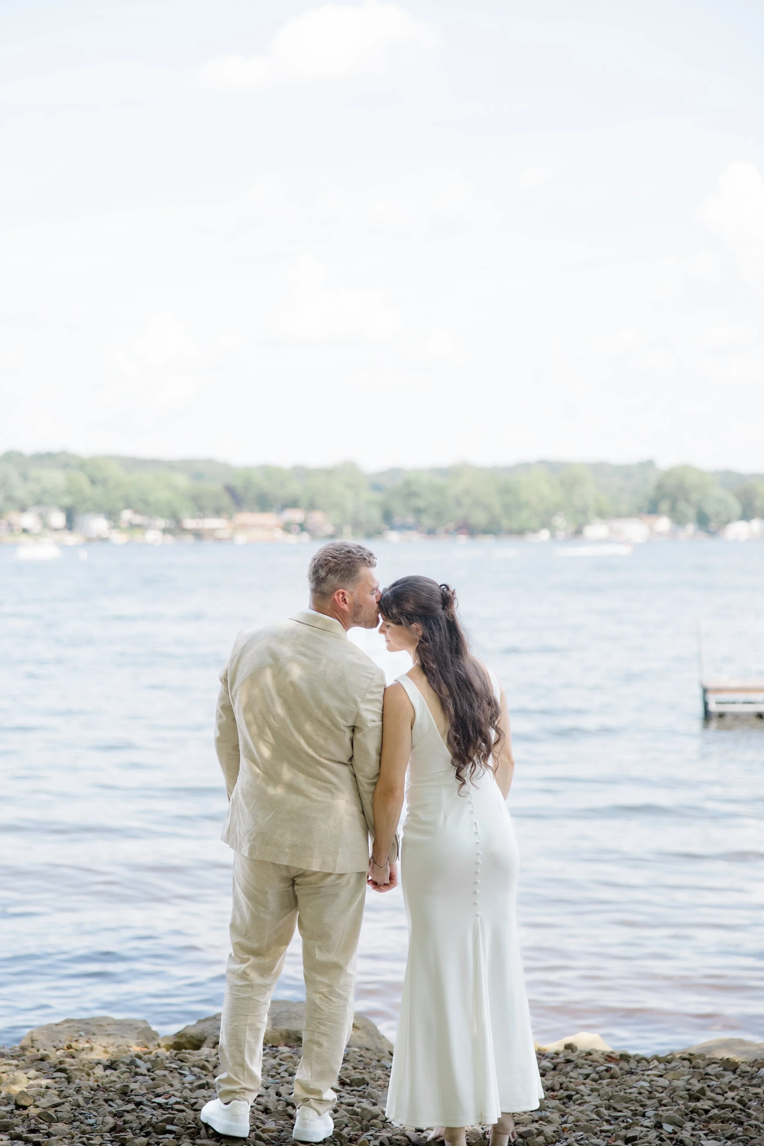 A couple in wedding attire holding hands and sharing a kiss by the water at a lakeside, with trees and boats in the background.