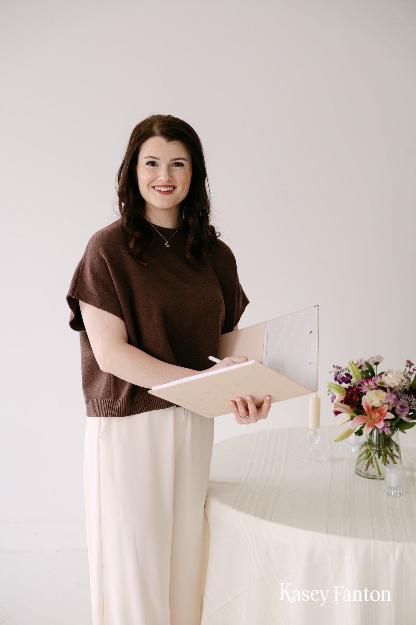 An Indianapolis event planner with dark hair is standing next to a table with a flower arrangement, holding a notebook and a pen, smiling at the camera.