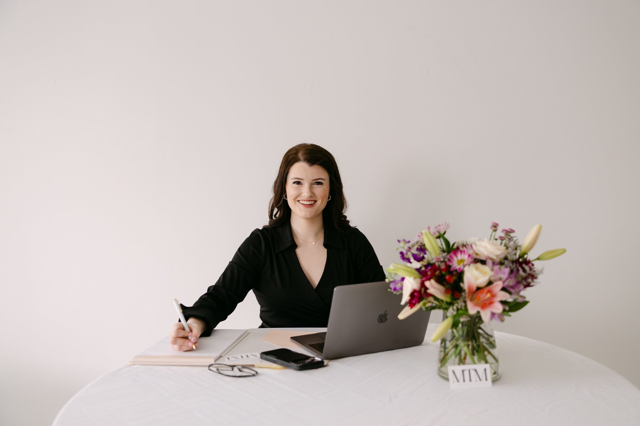 A woman with brown hair and a black blouse sitting at a table with a laptop, bouquet of flowers, notebook, and phone, smiling at the camera.