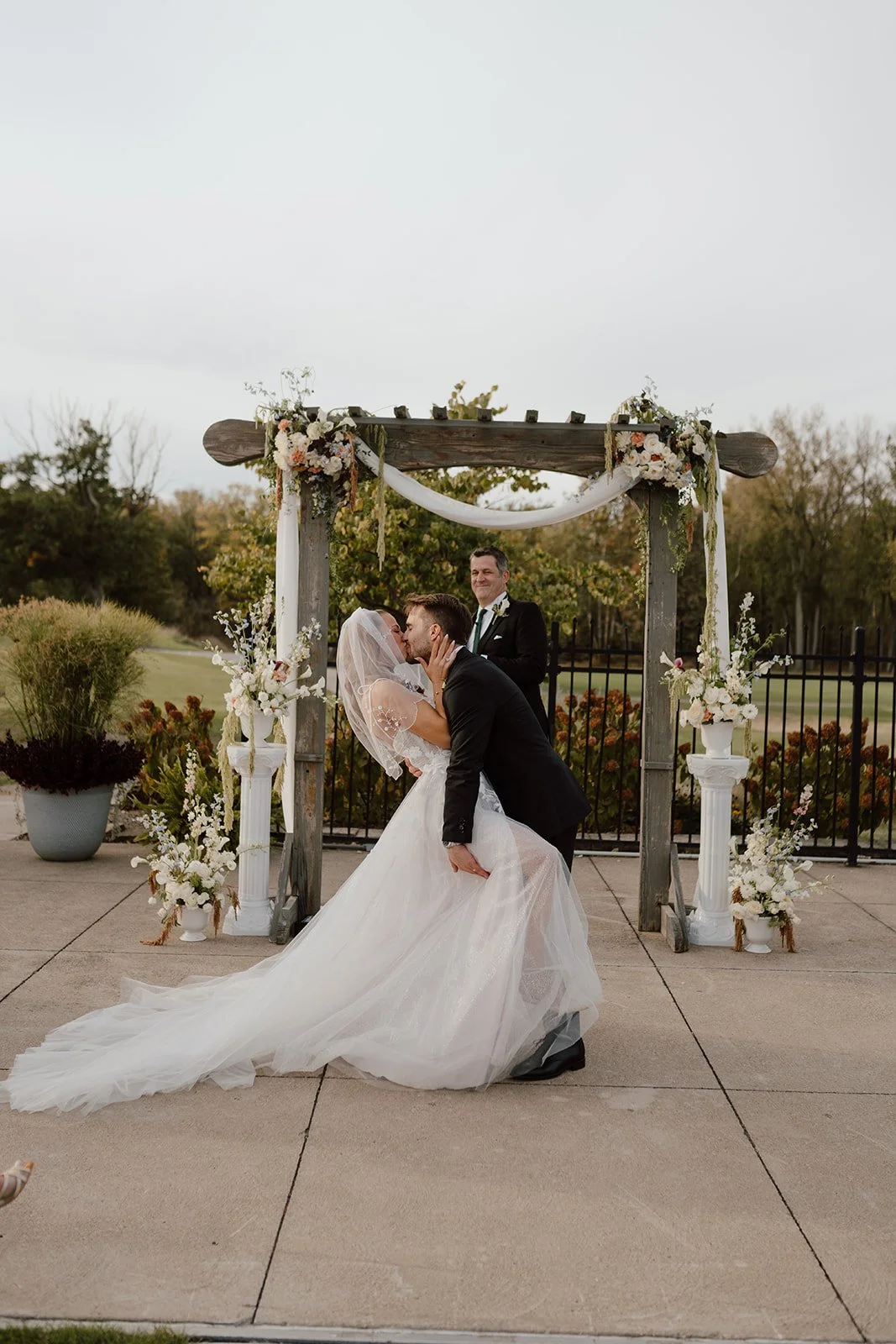 A bride and groom share a kiss during their wedding ceremony, with a officiant smiling in the background. They are standing under a rustic wooden wedding arch decorated with white and blush flowers and draped fabric, outdoors on a paved patio with some greenery and trees in the background.