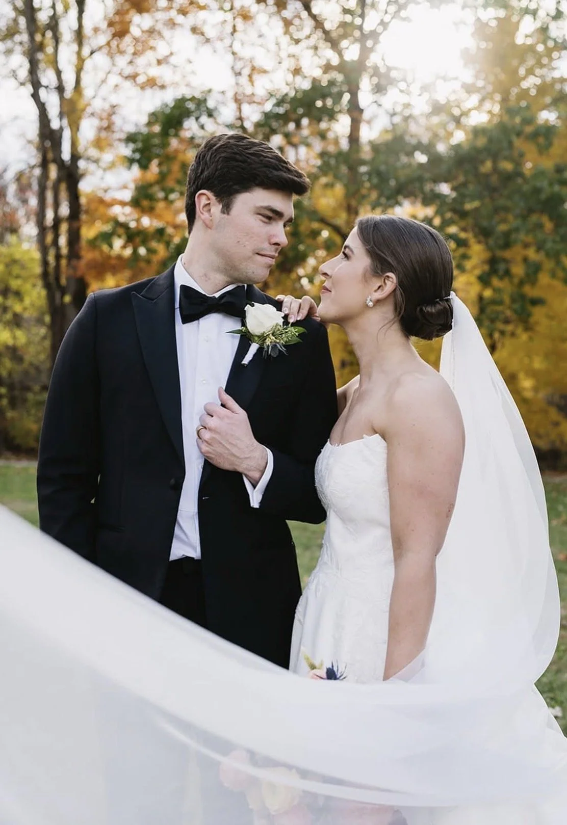 A bride and groom in wedding attire standing close outdoors during fall, with trees displaying autumn colors in the background. The groom is in a black tuxedo with a bow tie and boutonniere, while the bride is in a strapless white wedding gown with a veil and pearl earrings.
