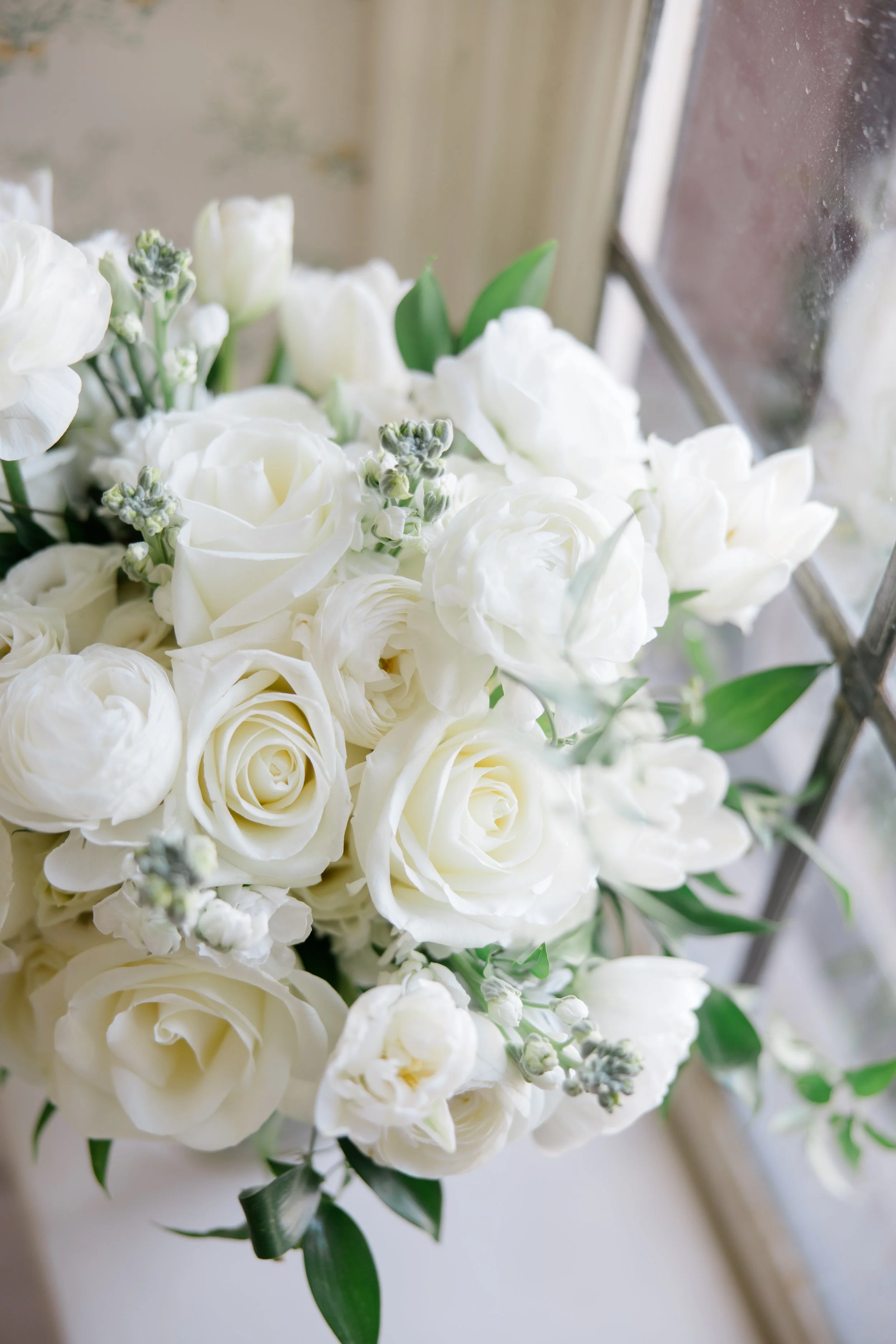 A bouquet of white roses, peonies, and small buds sitting near a window.