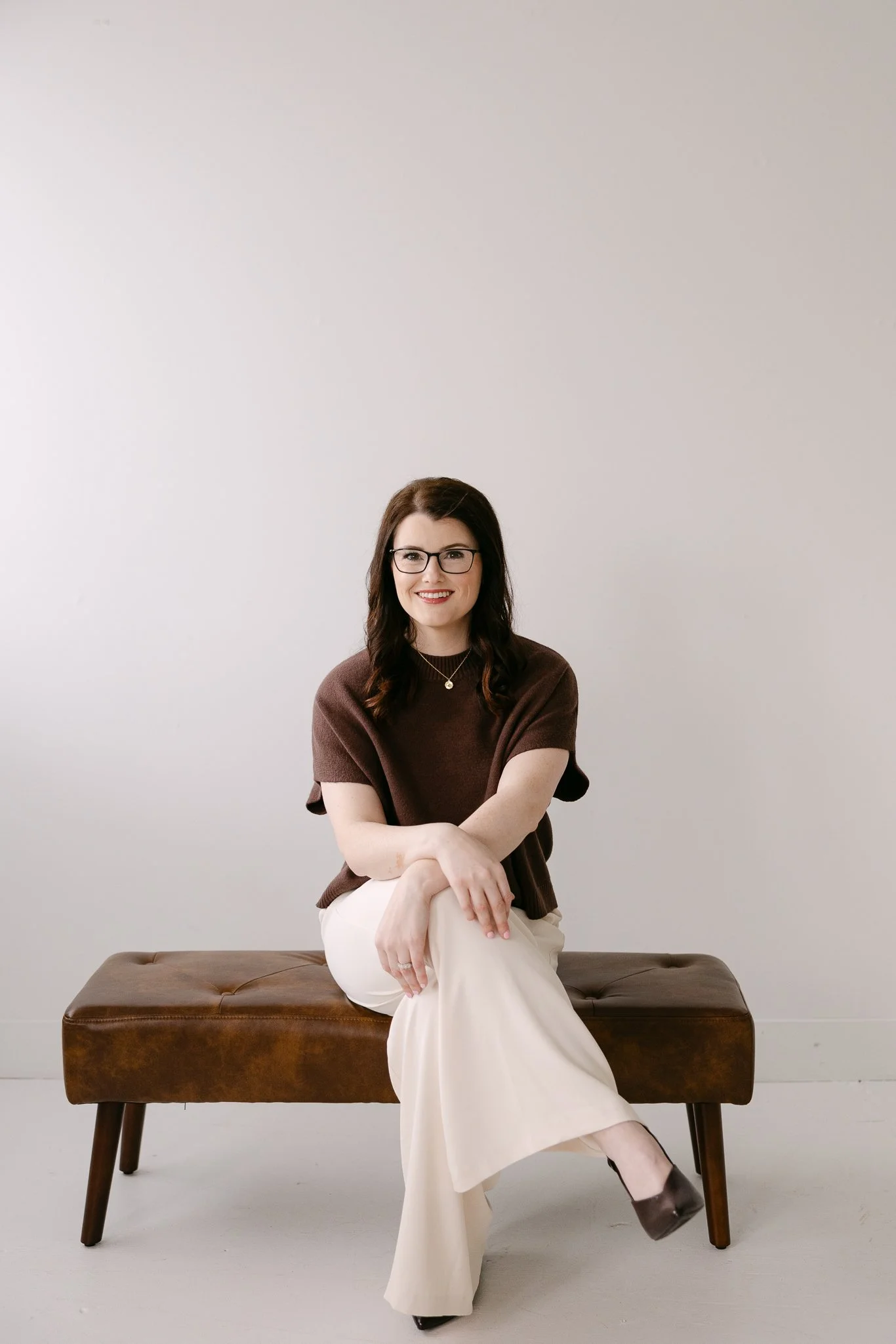 An Indiana wedding planner sitting on a brown leather bench in front of a plain light-colored wall. She has dark hair, glasses, and is smiling. She wears a brown top, off-white pants, and dark shoes.