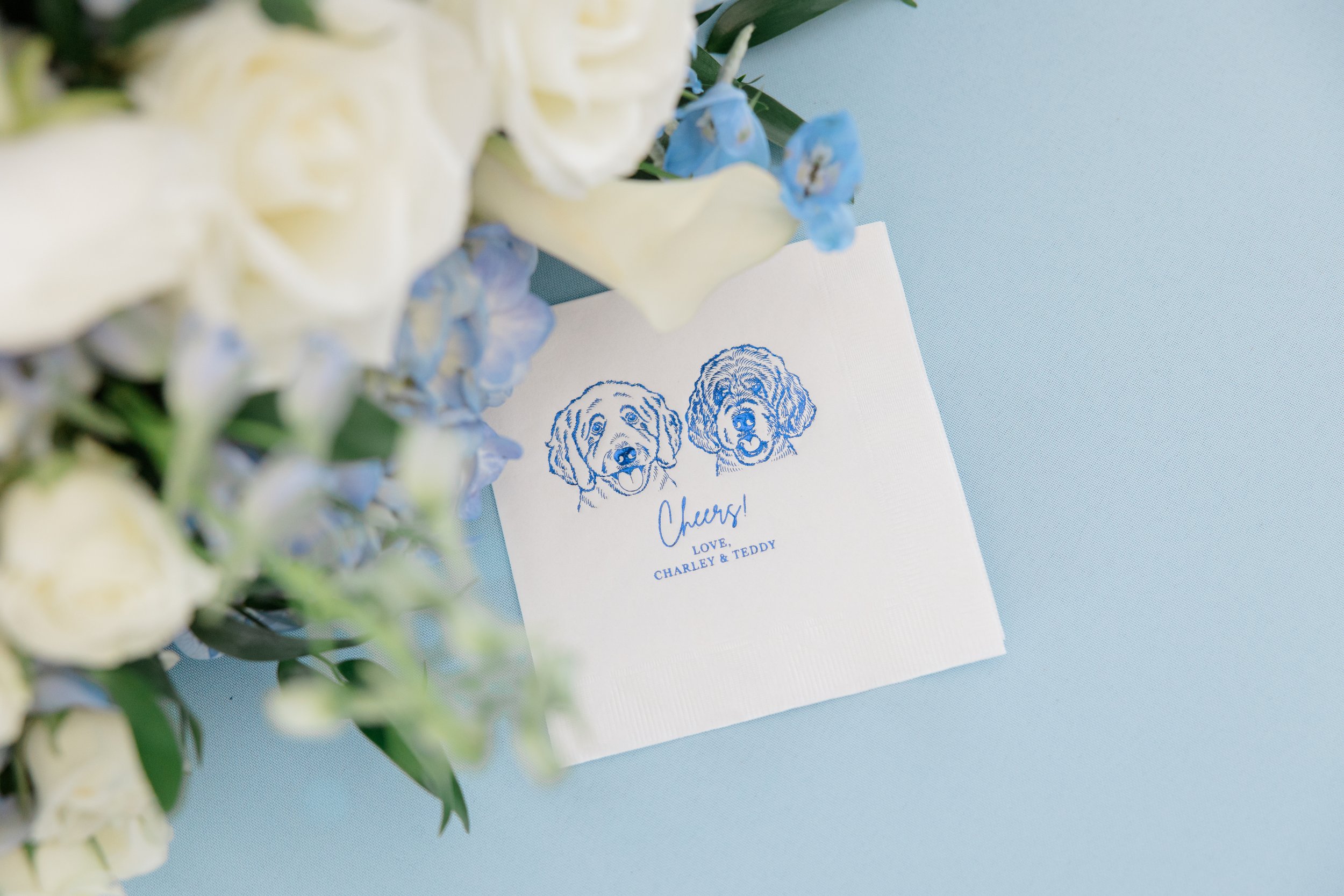 A floral arrangement with white and blue flowers next to a white card featuring blue line-drawings of two dogs, with the word 'Cheers!', and a message 'Love, Charley & Teddy'.