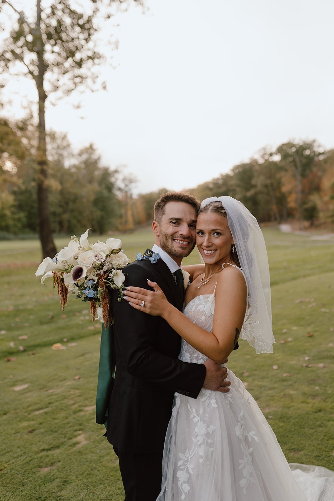 A happy couple in wedding attire embracing outdoors on a grassy field with trees in the background, the bride holding a bouquet of white and purple flowers.