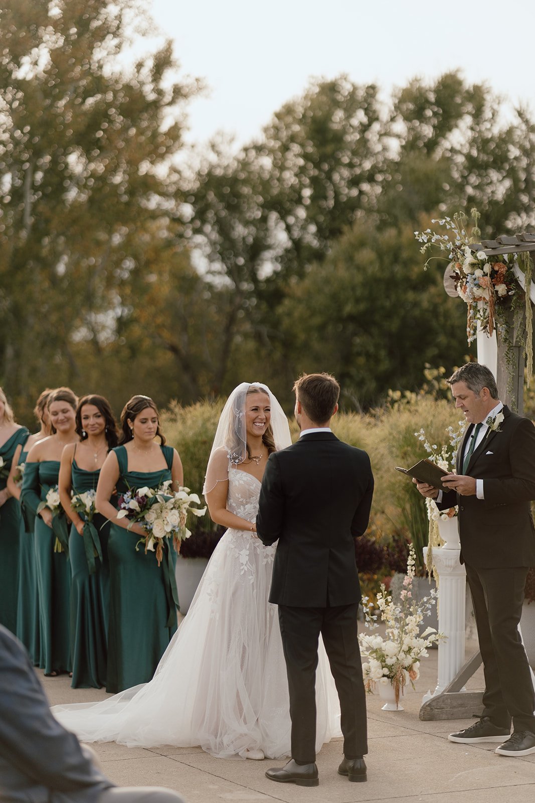 A bride and groom exchanging vows during a wedding ceremony outdoors, with bridesmaids in green dresses standing behind them, and an officiant reading from a book.