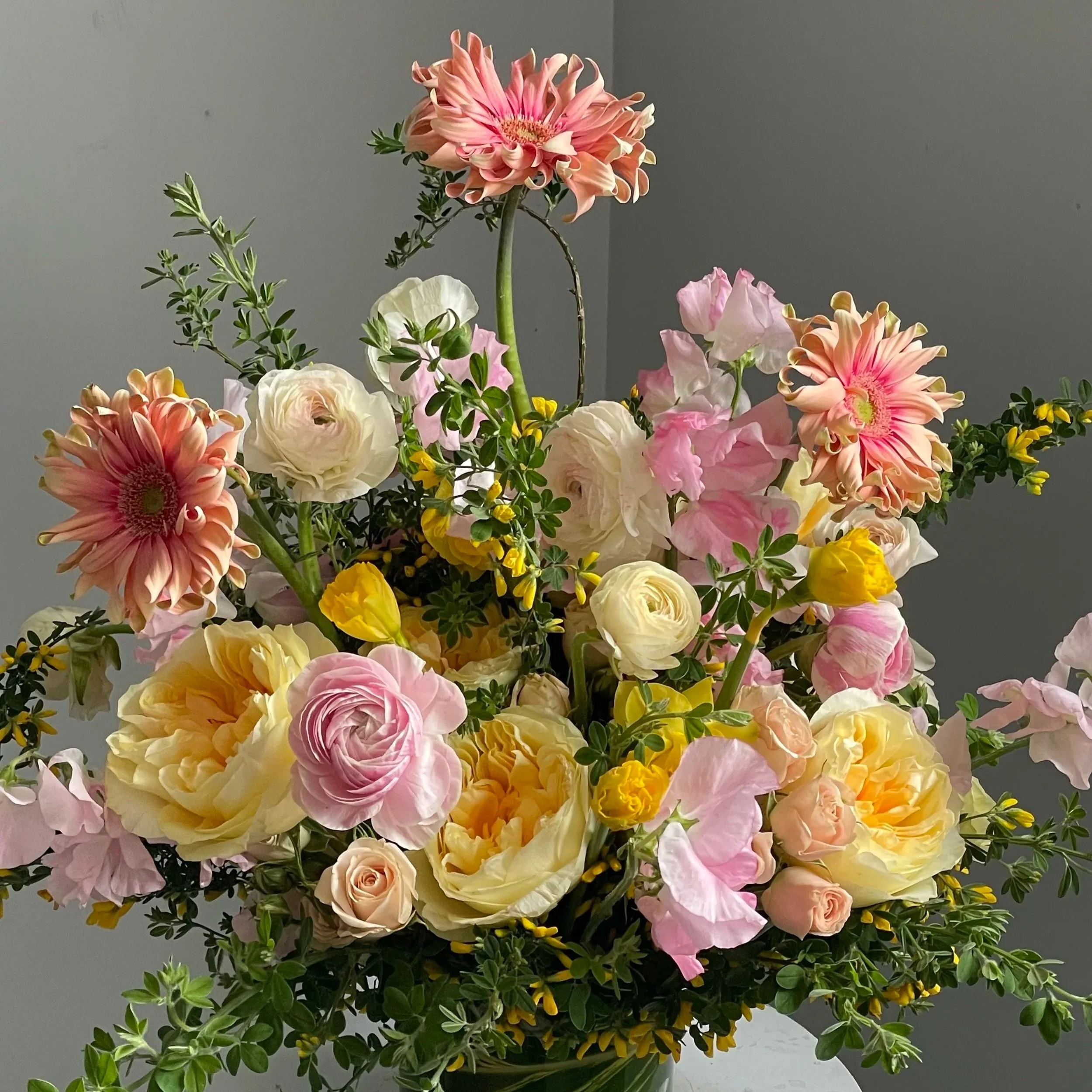 A colorful bouquet of various flowers including pink, white, yellow, and peach blossoms arranged in a vase against a gray background.