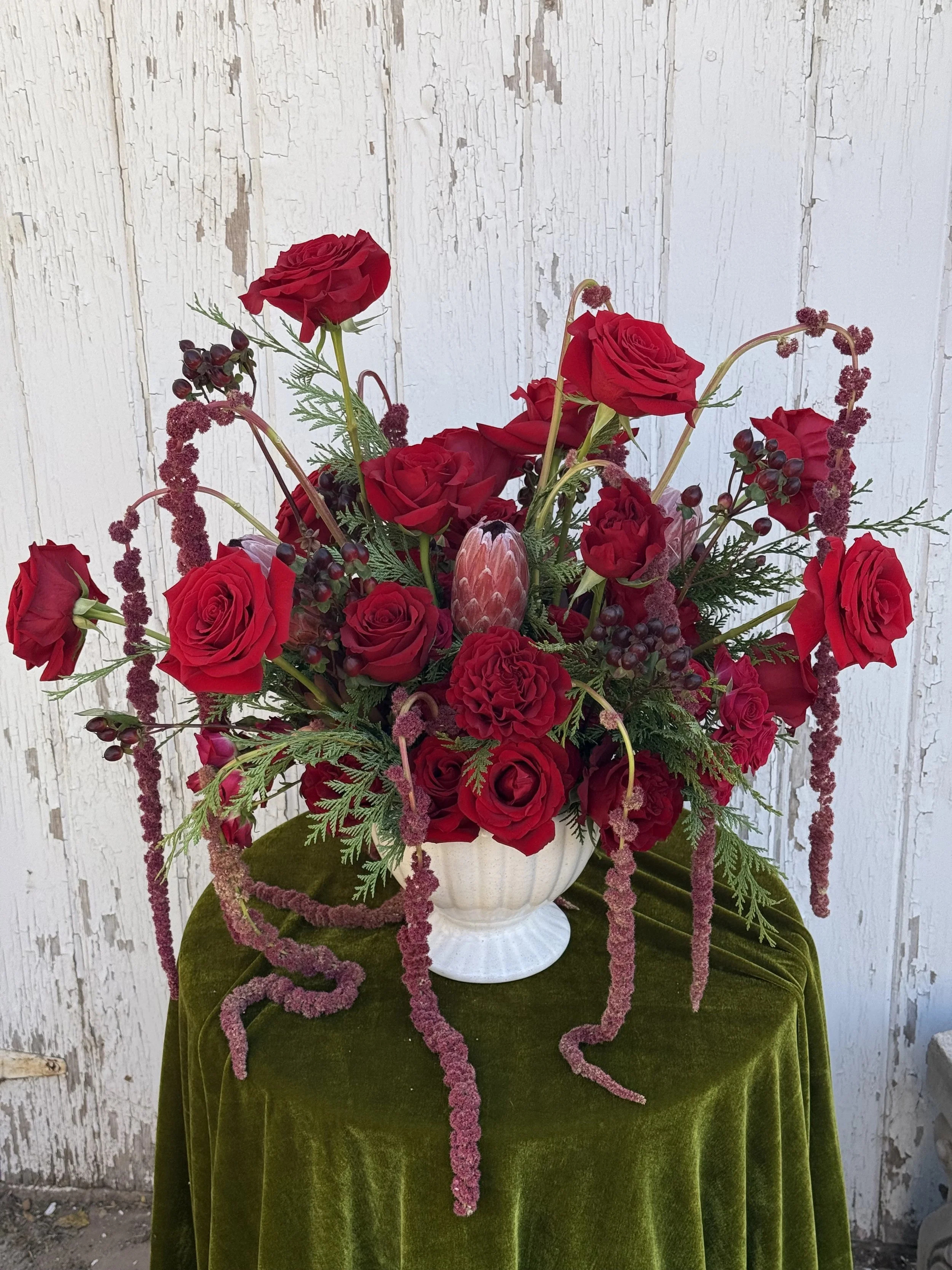 A floral arrangement featuring red roses, pink protea, purple berries, and greenery in a white vase, placed on a green velvet-covered table against a distressed white wooden wall.