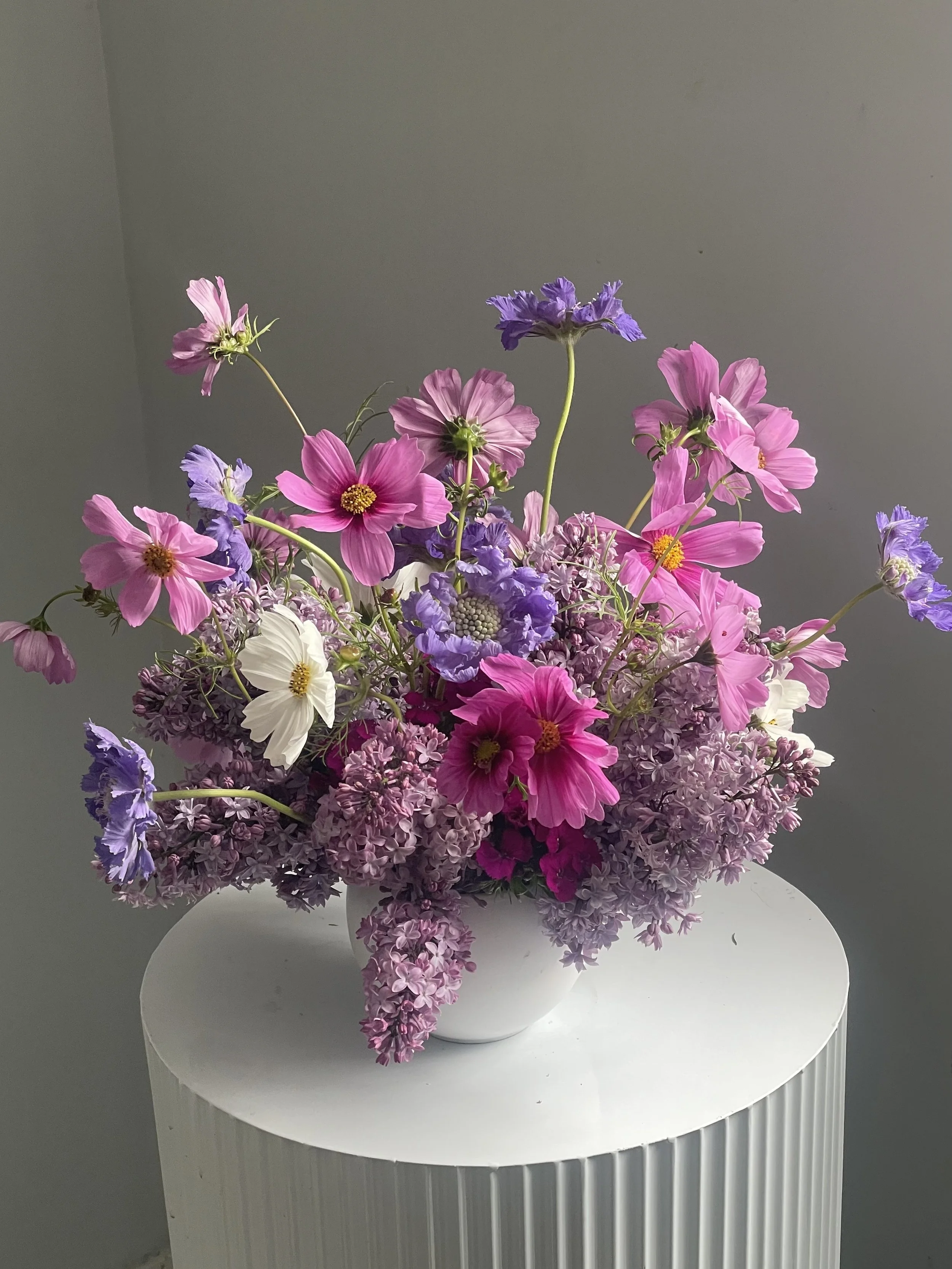 A white vase filled with pink, purple, white, and lilac flowers placed on a white round table.
