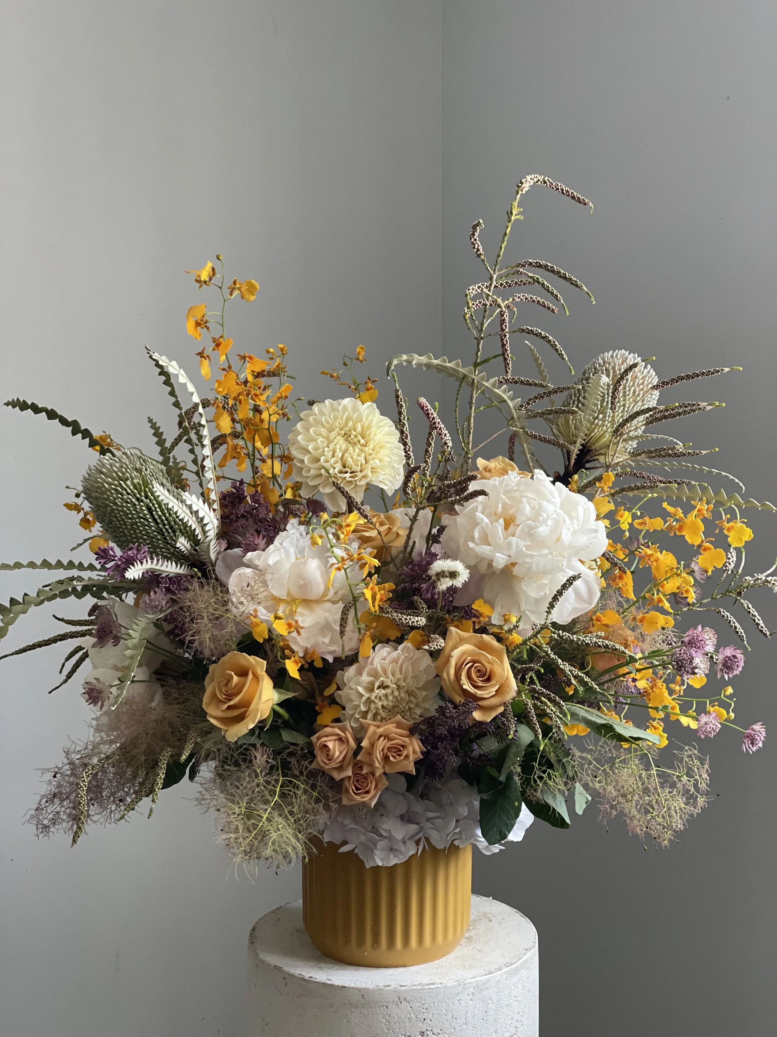 A bouquet of various flowers including roses, dahlias, and yellow blooms in a yellow vase placed on a white pedestal against a plain gray background.