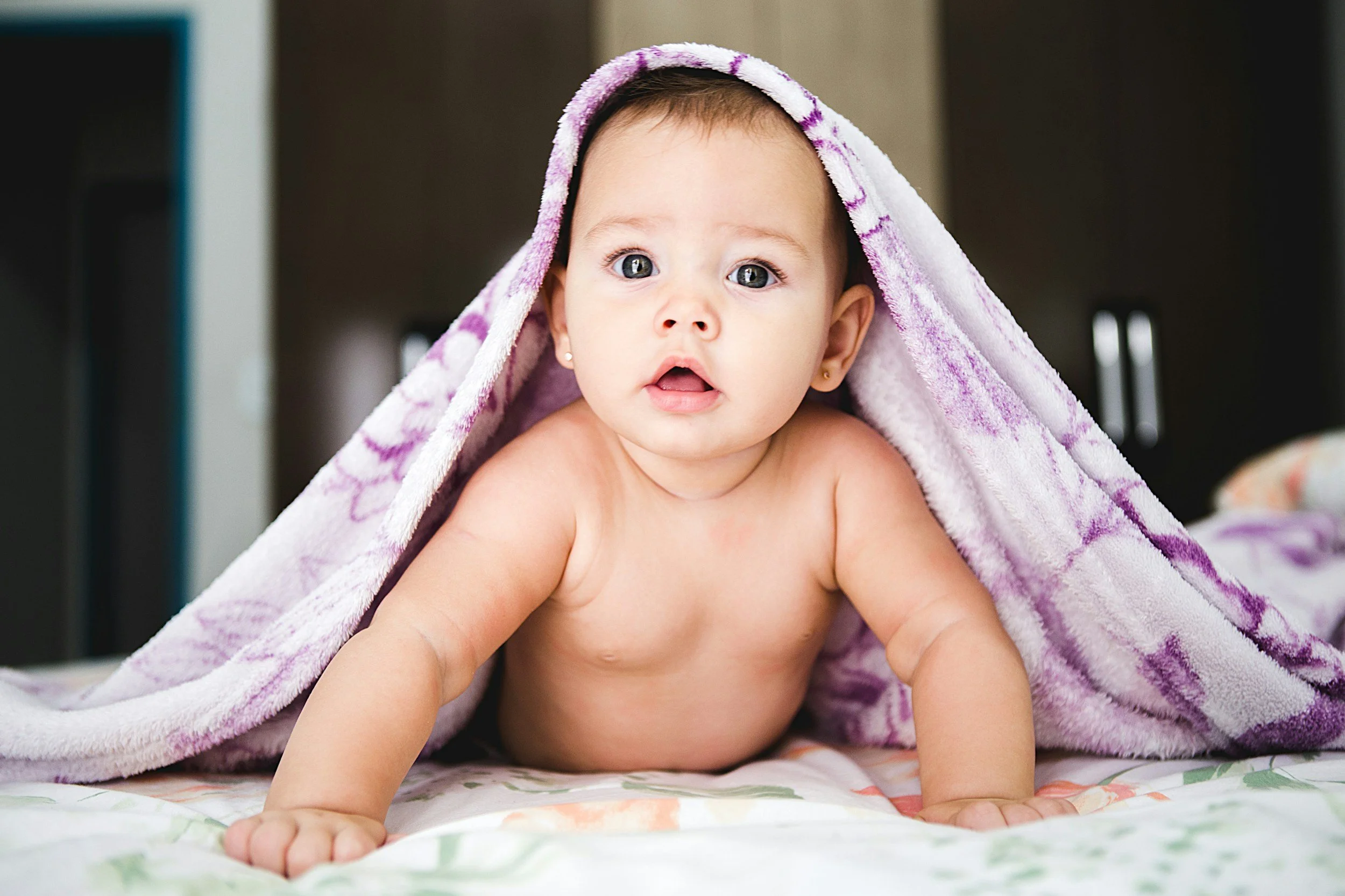 A baby crawling on a bed, partially covered with a purple and white towel draped over their head.