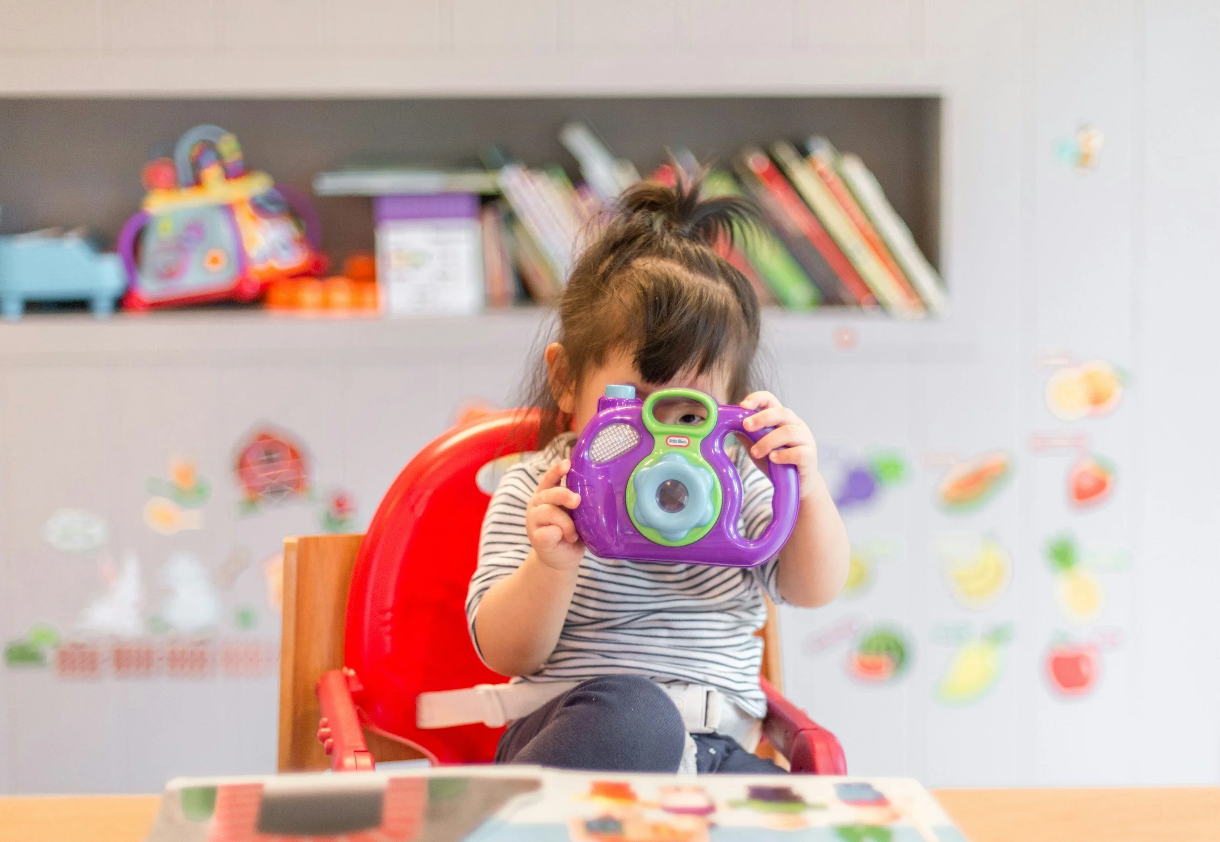 A young girl sitting in a red high chair, holding a colorful toy camera in front of her face. The background features a bookshelf and wall decorations with various stickers.