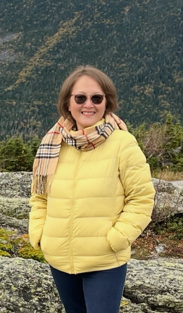 A woman in a yellow puffy jacket and sunglasses standing outdoors in front of a mountainous landscape with rocks and trees.